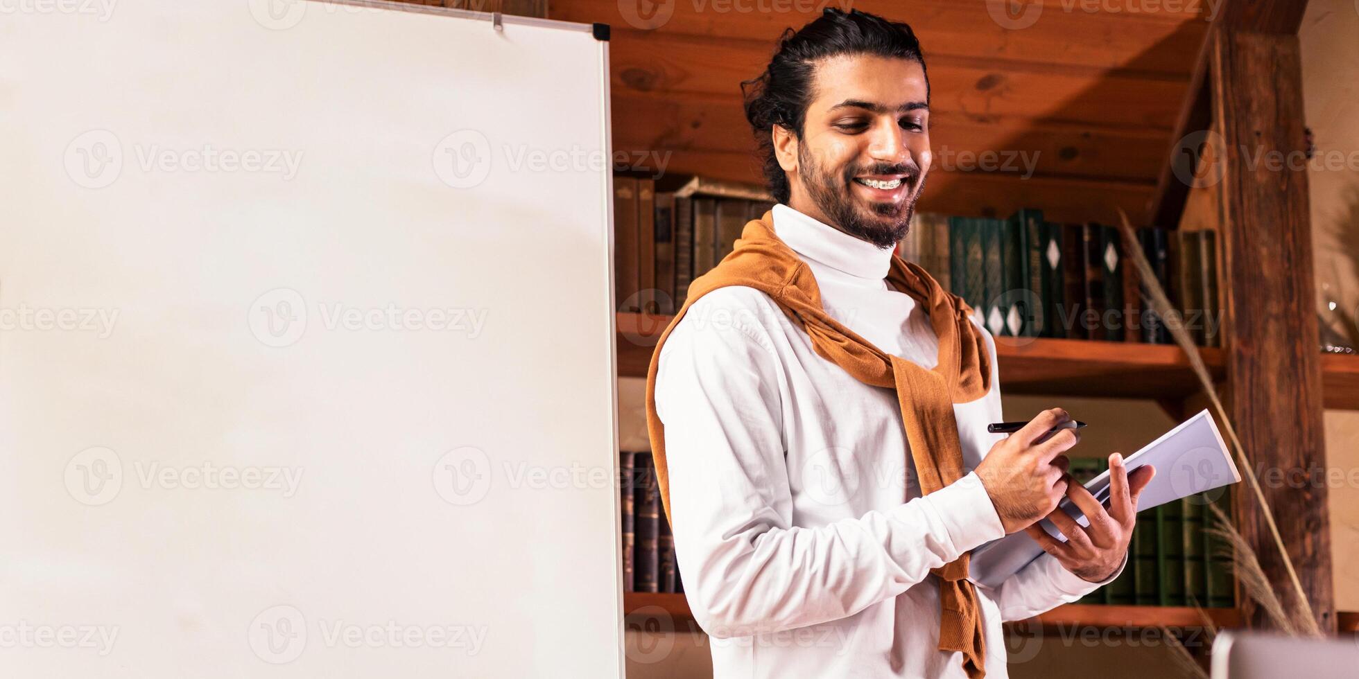 Distance Teaching. Cheerful Indian Teacher Having English Class Online Talking To Laptop Webcam Standing Near Blackboard And Explaining Language Grammar Rules At Home. E-Teaching And E-Learning photo
