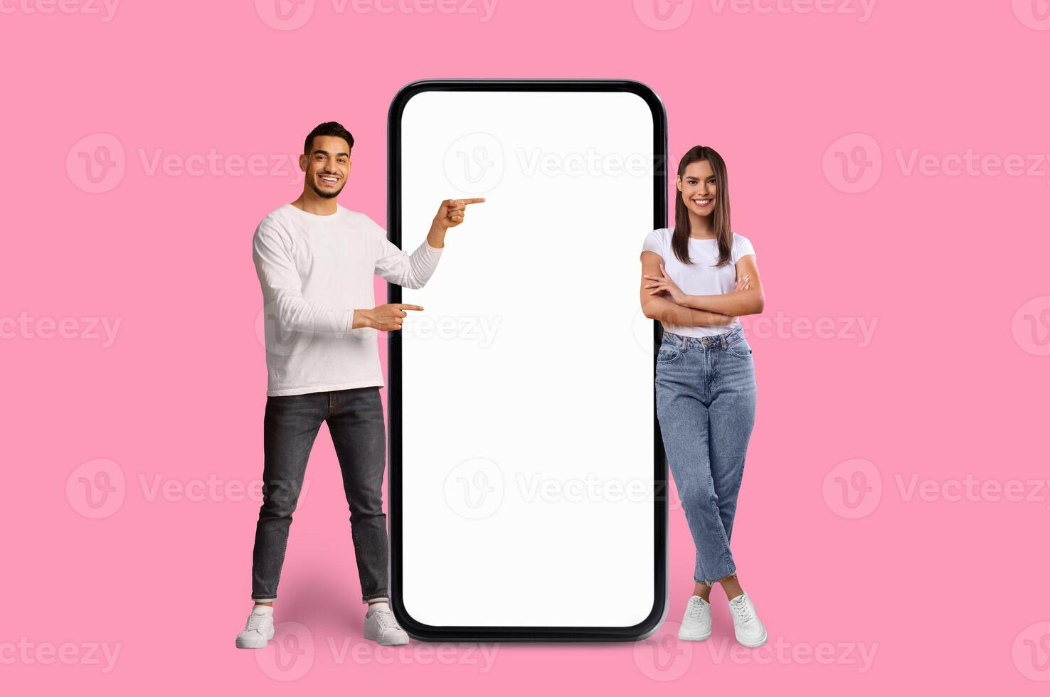 A cheerful Multiracial young couple stands next to a giant smartphone pointing and smiling at its blank screen photo