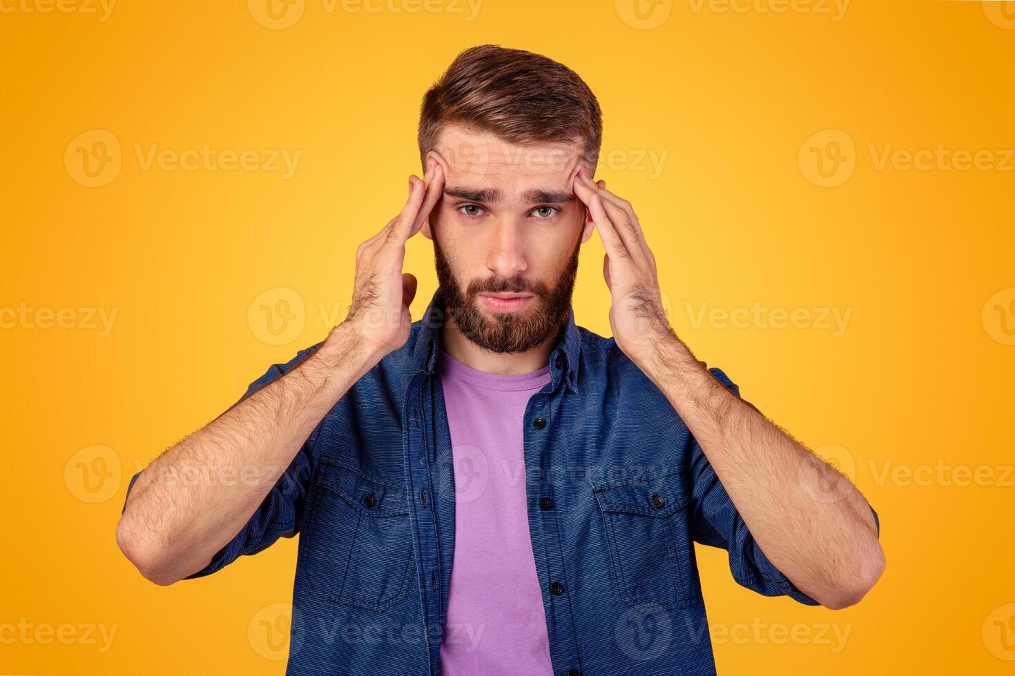 Millennial guy suffering from headache, massaging temples to get rid of pain on orange studio background. Young man having hypertension, feeling stressed or exhausted, cannot find problem solution photo