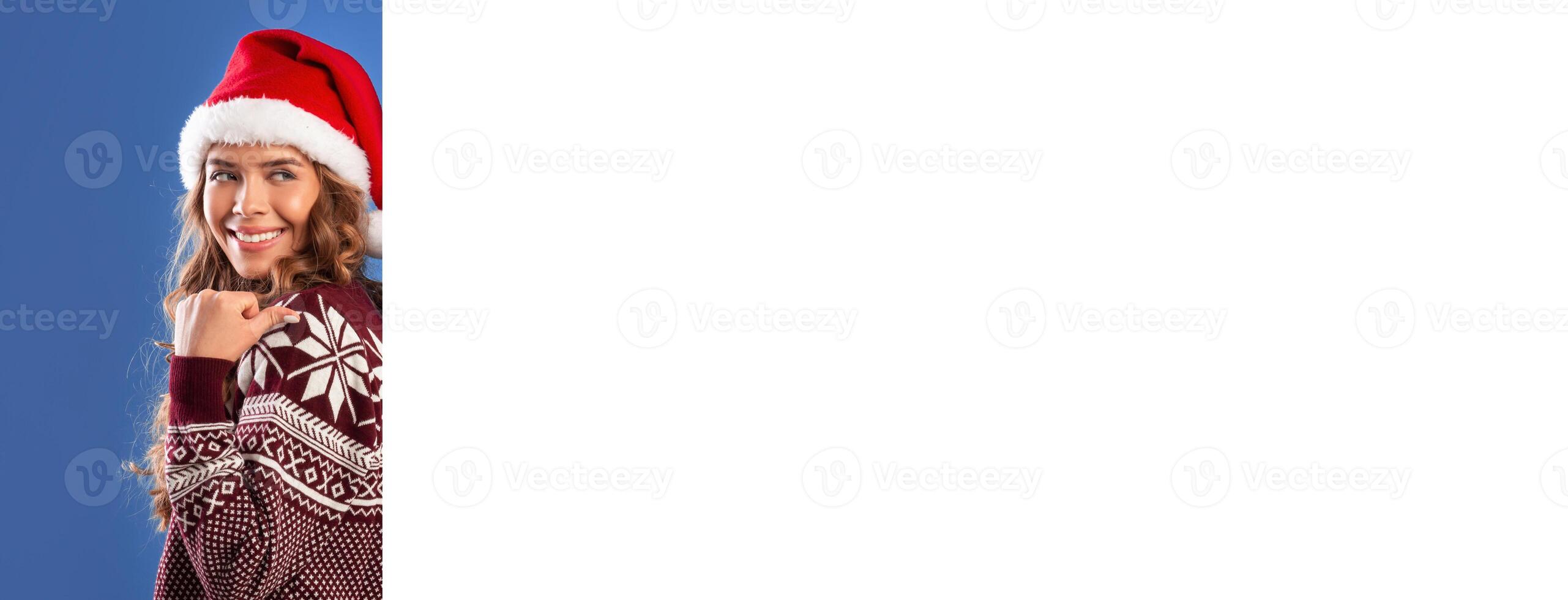 Positive young lady in warm sweater and Santa hat pointing at empty banner with space for your ad on blue background, mockup for design. Panorama. Lovely lady making Christmas promo photo