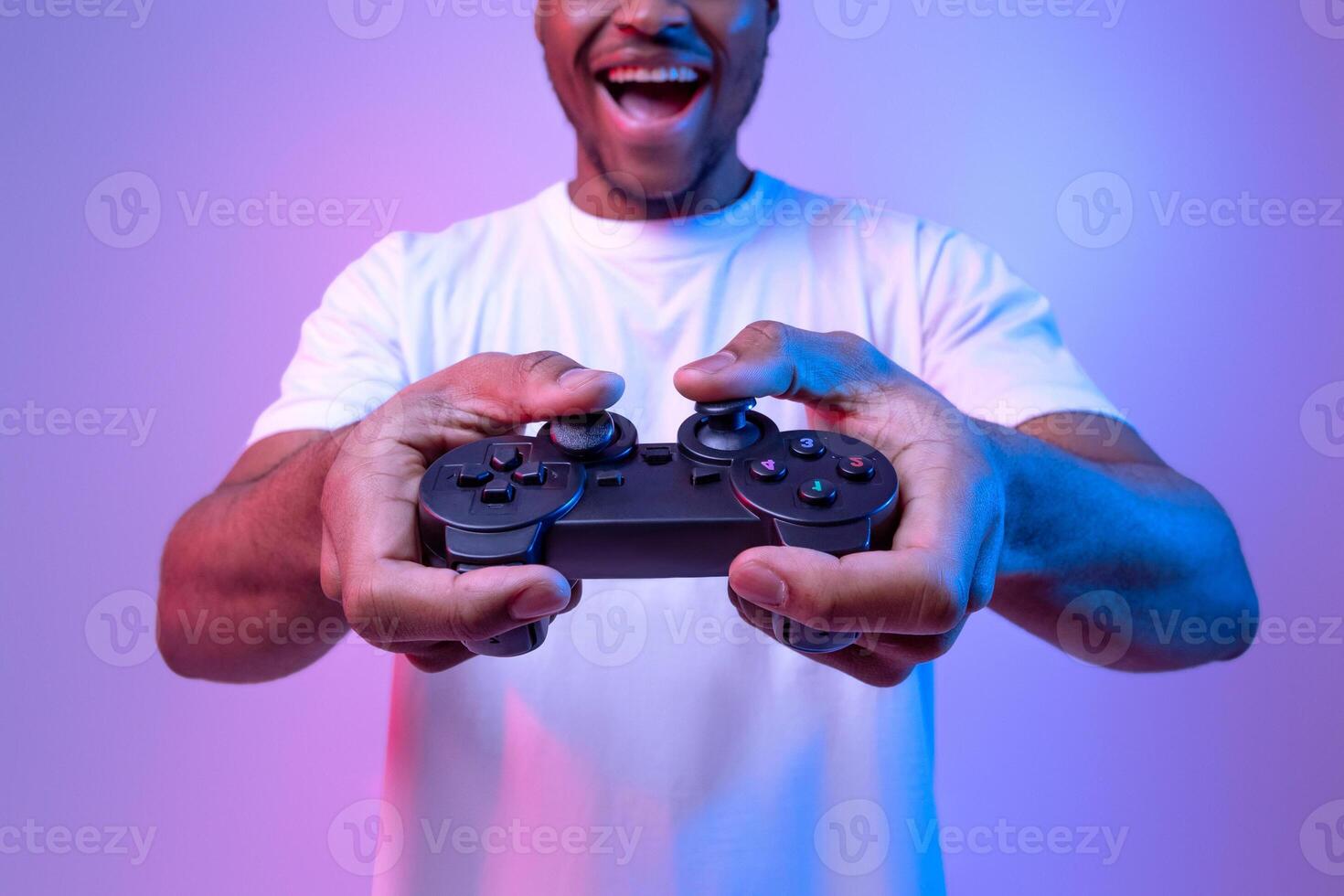 Excited african american guy playing games with joystick, joyful black man enjoying modern entertainment, having fun in vivid neon light on purple studio background, selective focus, copy space photo