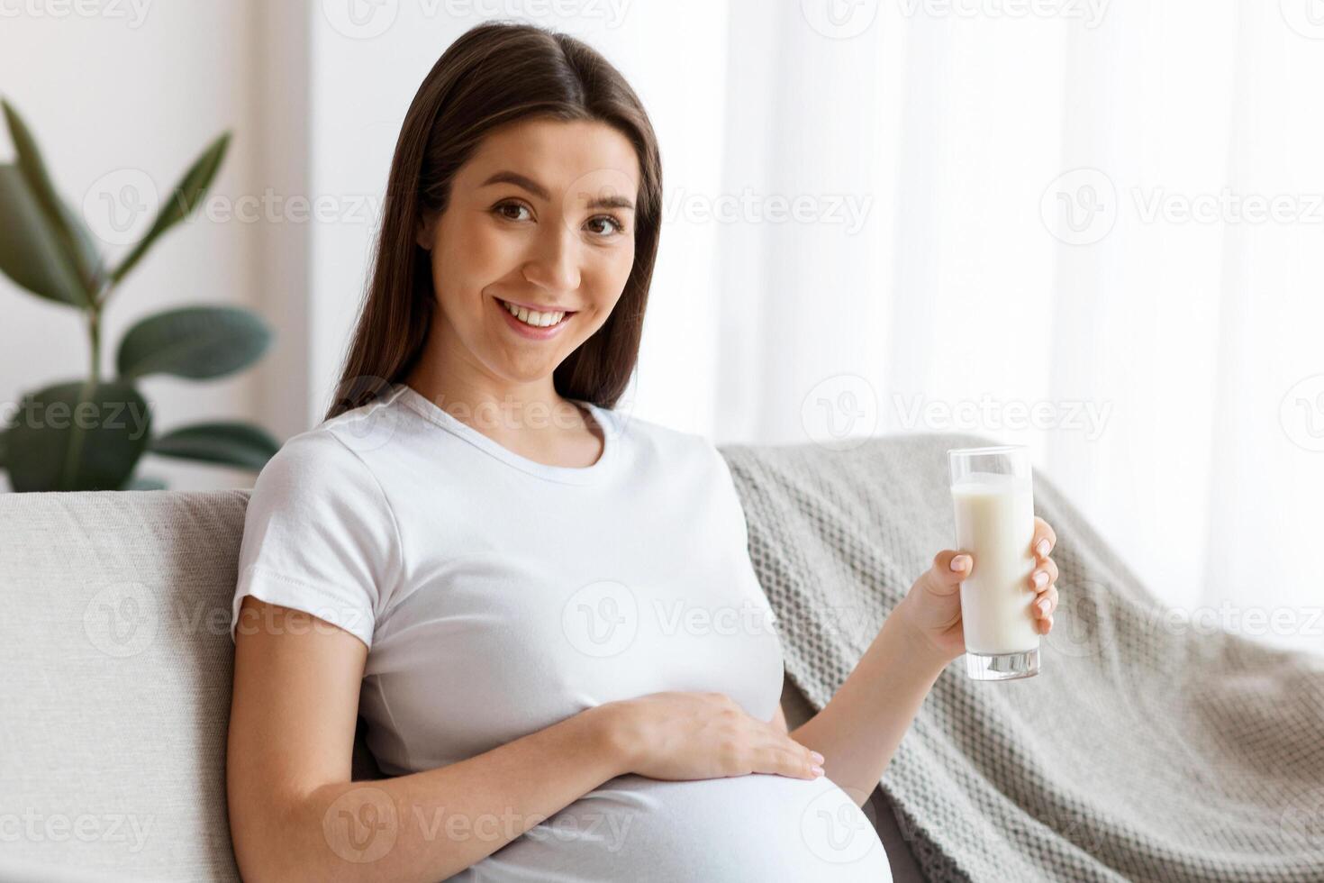 Pregnancy Nutrition Concept. Portrait Of Happy Young Pregnant Woman With Glass Of Milk In Hand Relaxing On Comfortable Couch In Living Room, Beautiful Expectant Lady Enjoying Calcium Drink photo