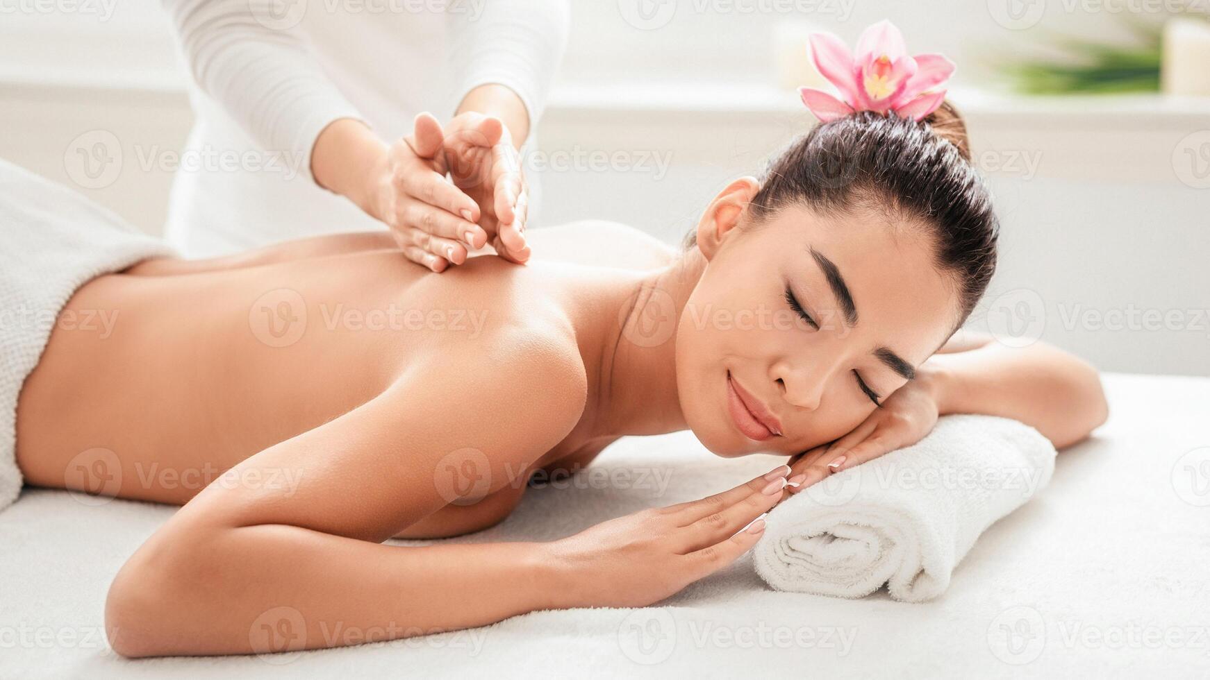 Young Asian Lady Getting Back Massage Treatment From Professional Masseur In Spa, Attractive Korean Woman Lying Naked On Table With Rolled Towel Under Head, Enjoying Relaxing Beauty Procedures photo