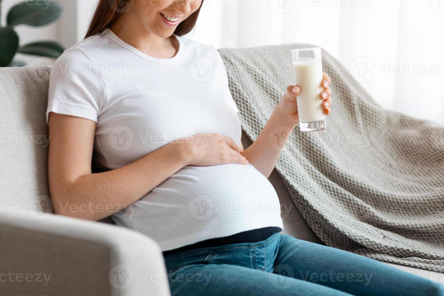 Healthy Calcium Drink. Smiling Pregnant Lady Holding Glass Of Milk, Sitting On Couch At Home And Embracing Her Belly, Young Expectant Woman Resting In Living Room, Enjoying Pregnancy Time, Free Space photo