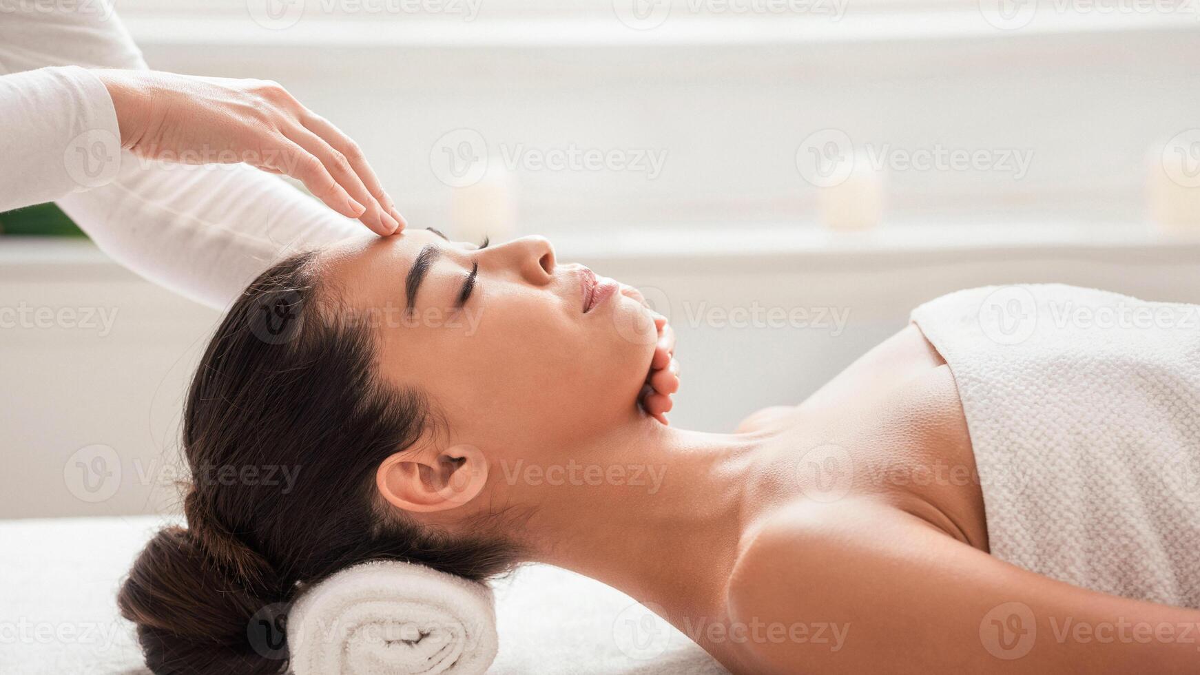 Masseur doing acupressure massage to young asian woman, pressing on her forehead. Relaxed korean lady enjoying wellness day at spa salon, having beauty treatments at cosmetology cabinet, side view photo