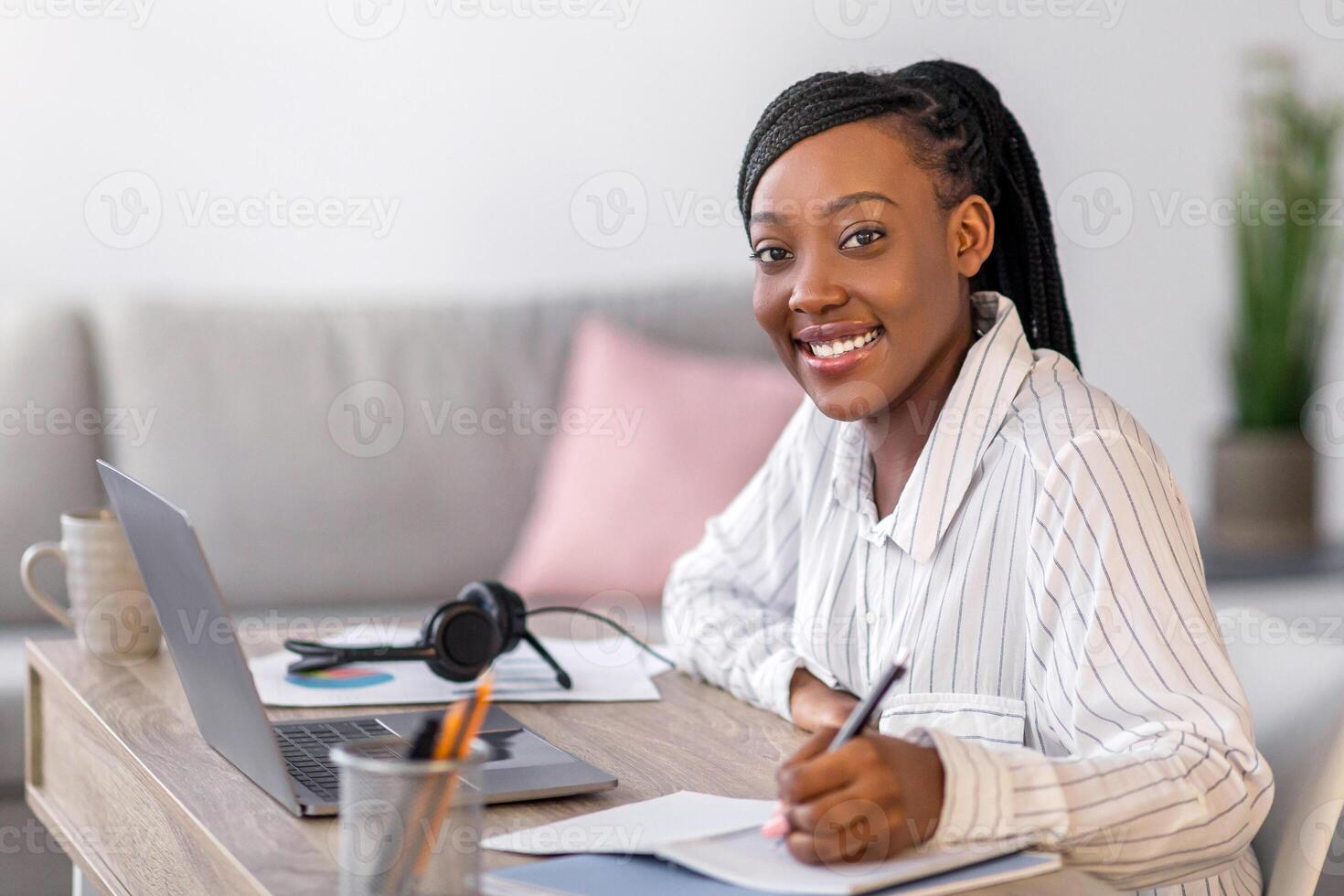 Pretty young black woman enjoying online course, smiling african american lady sitting at workdesk in front of laptop, taking notes in notepad, home interior, side view, copy space photo