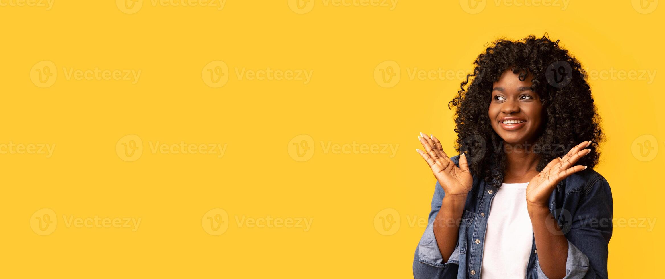 Emotional african american woman with beautiful curly hair looking at empty space, holding hands in surprised gesture and smiling, yellow studio background, panorama. Amazed african american lady photo