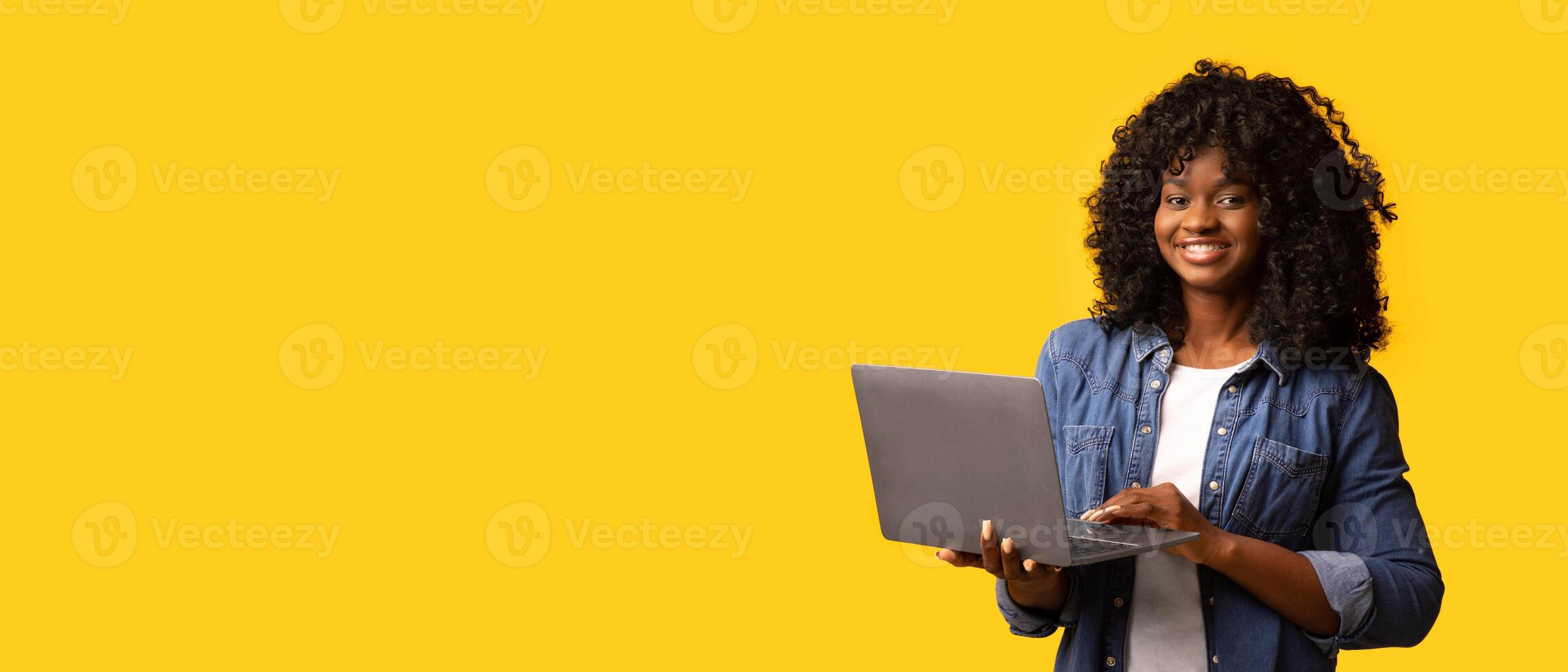 Happy african american young woman holding newest laptop and smiling, yellow studio background, panorama with copy space. Cheerful black lady using notebook, shopping online or websurfing photo