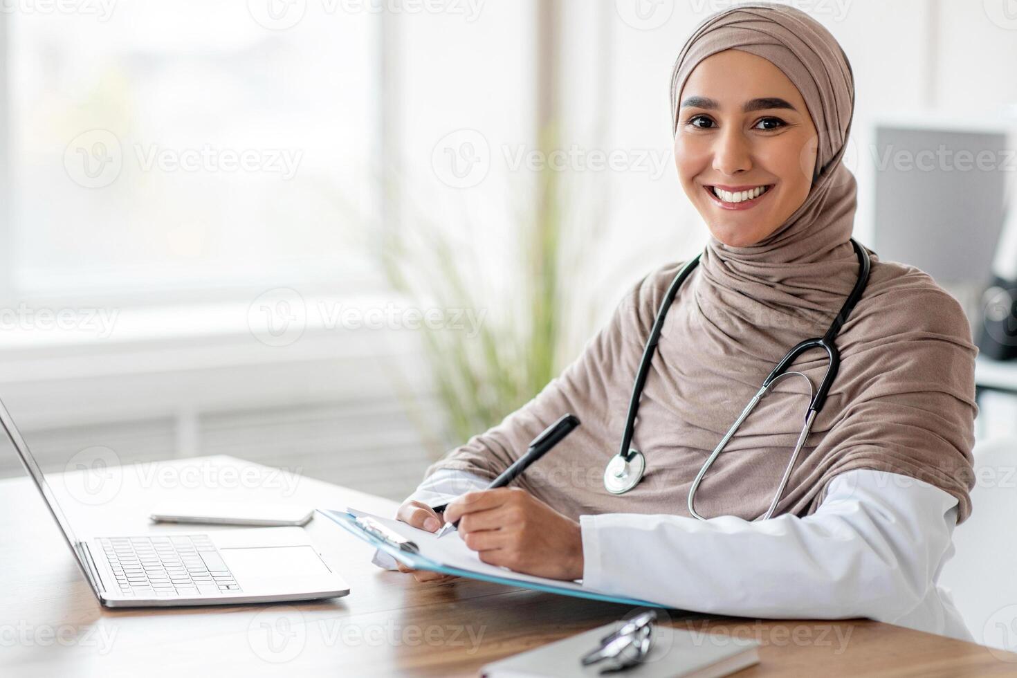 Smiling muslim woman doctor in headscarf taking notes in medical chart, sitting at workdesk with laptop at her cabinet in clinic, taking anamnesis while having conversation with patient, free space photo