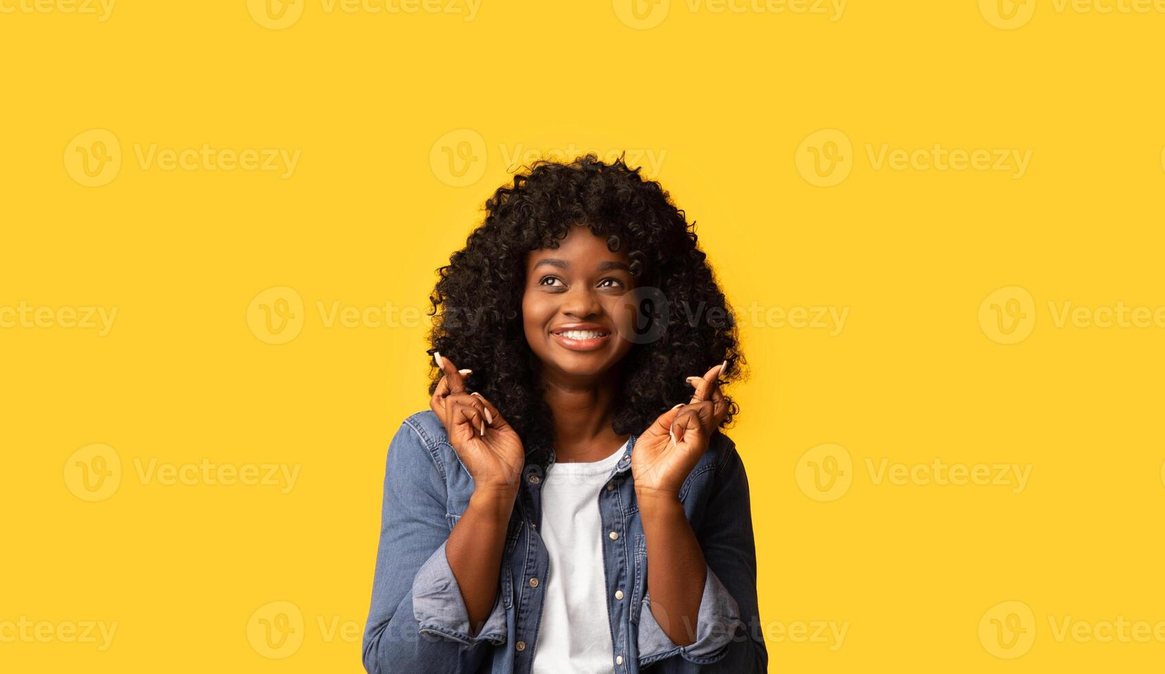 Wish, desire, superstition concept. Happy african american young lady with crossed fingers looking aside on yellow studio background. Superstitious black woman making wish, having hope, copy space photo