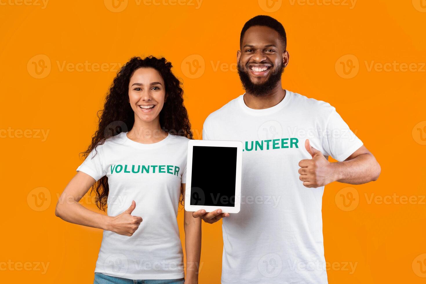 Couple Of Multiracial Volunteers Holding Tablet Computer With Empty Screen Gesturing Thumbs Up Standing On Yellow Background, Smiling To Camera. Studio Shot, Mockup photo