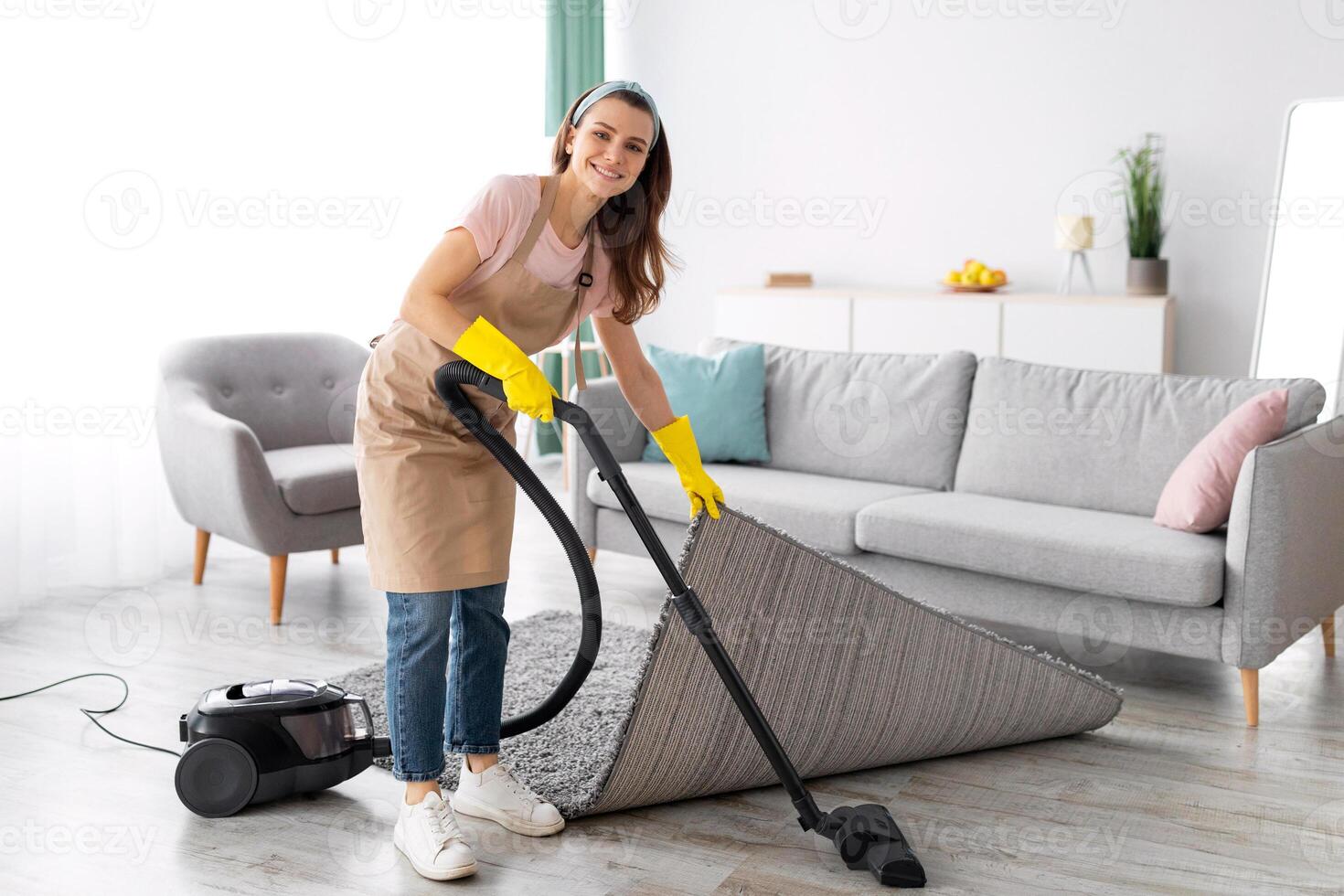 Positive female housekeeper using vacuum cleaner to tidy modern apartment. Happy young woman doing domestic chores, keeping her house tidy. Sanitary service concept photo