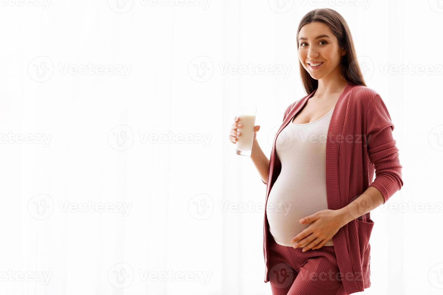 Healthy Drinks During Pregnancy. Portrait Of Smiling Young Pregnant Woman Holding Glass With Milk While Standing Near Window At Home, Happy Expectant Lady Enjoying Calcium Drink, Copy Space photo