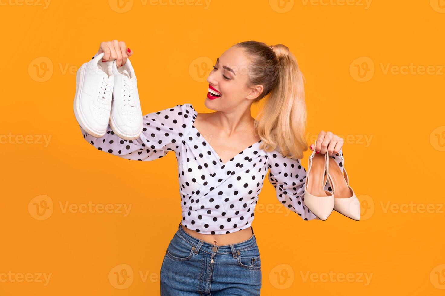 Making Decision. Portrait of smiling lady holding pair of high heels shoes and white sneakers in hands, can't choose between footwear and style, isolated over orange studio background photo