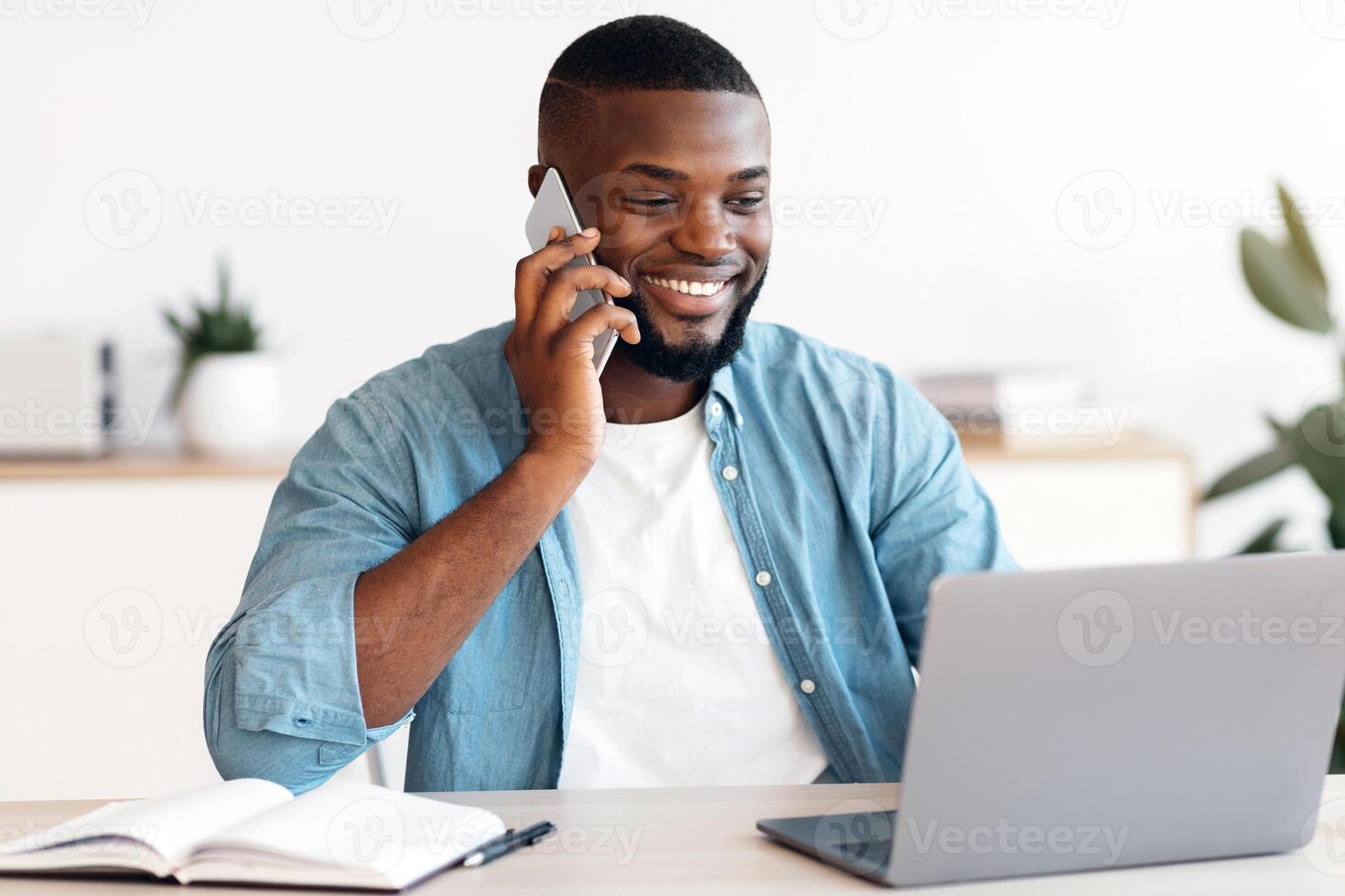 Remote Job. Black Freelancer Guy Using Cellphone And Laptop At Home Office, Smiling African American Man Sitting At Desk, Working On Computer And Enjoying Phone Conversation, Free Space photo