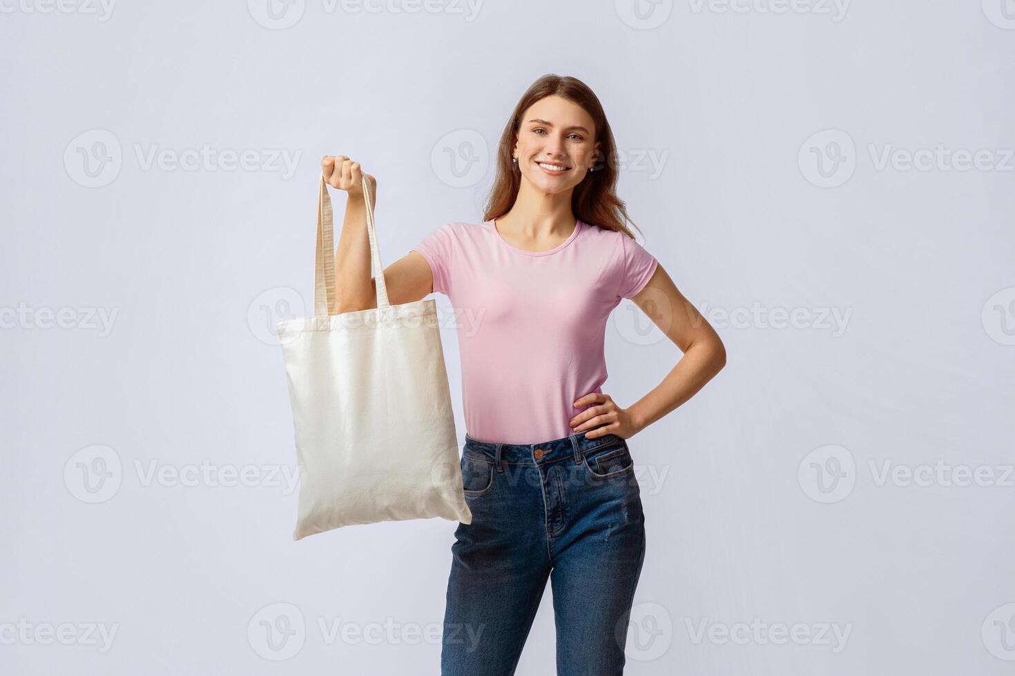 Smiling Millennial Woman Posing With Blank Eco Tote Canvas Bag, Demonstating Free Copy Space For Your Design Or Logo, Positive Young Lady Standing Over Light Studio Background, Looking At Camera photo