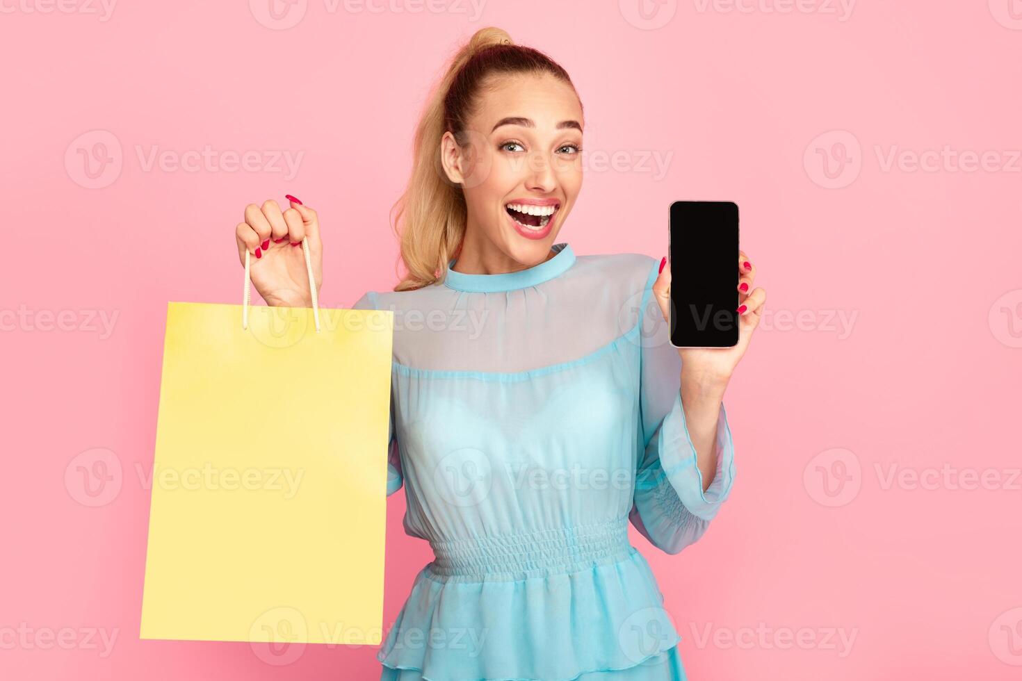 Shopping Application And Online Sales. Overjoyed woman showing mobile phone with black empty screen for mockup to camera and holding bag, standing and posing at pastel pink studio wall background photo
