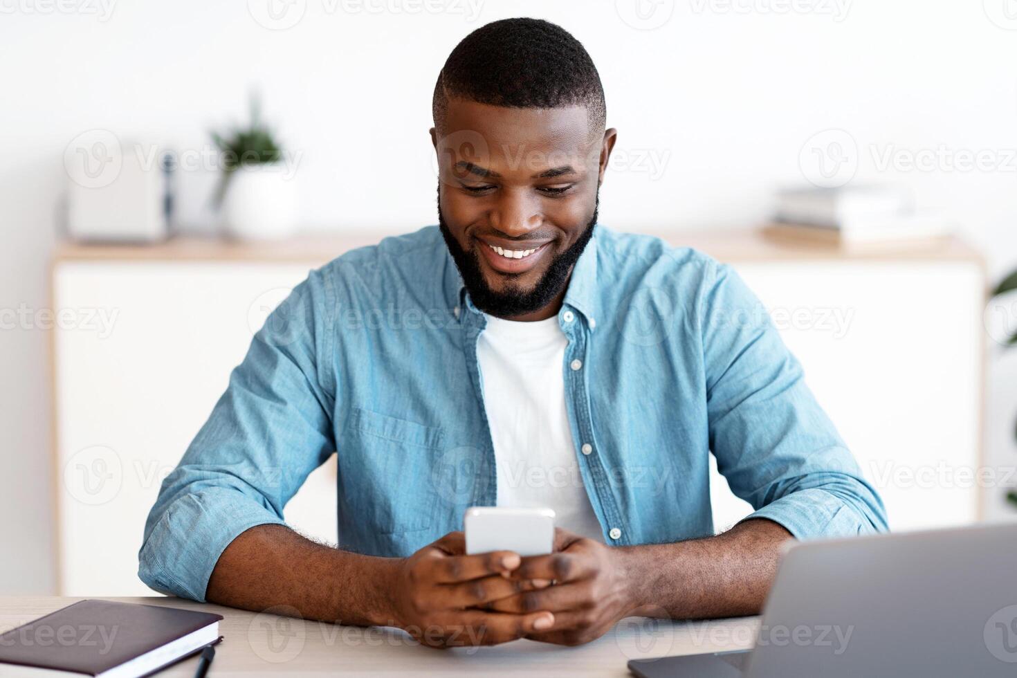 Young Black Freelancer Guy Sitting At Desk And Using Smartphone, Smiling Millennial African Man With Mobile Phone In Hands Messaging Or Shopping Online While Working At Home Office, Free Space photo