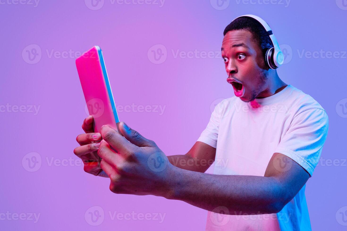 Online Offer. Shocked Black Guy In Wireless Headphones Looking At Digital Tablet With Amazement, Emotional African American Man Standing With Open Mouth Over Purple Background In Neon Light, Panorama photo