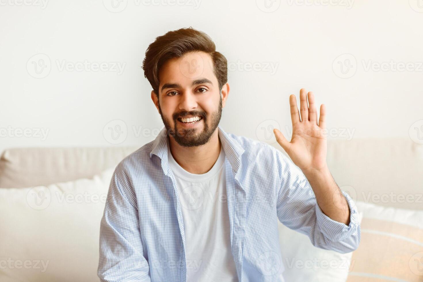 Greeting Concept. Portrait of friendly eastern man waving hello while sitting on couch at home, positive millennial arab guy smiling and looking at camera, saying hello, free space photo