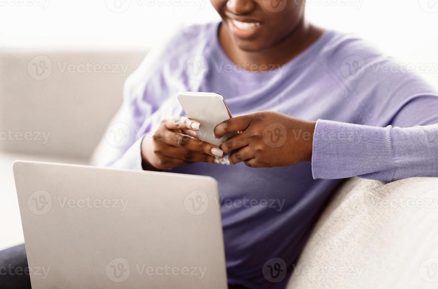 Scrolling News Feed. Closeup of unrecognizable african american smiling lady holding cellphone, typing sms message, sitting on couch with laptop. Black woman browsing internet, surfing web, using app photo