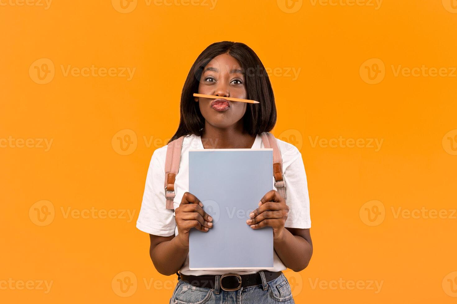 Funny young african american lady student with backpack, books and notebooks posing over orange studio background, placing pencil as moustache, copy space. Education, college, university concept photo