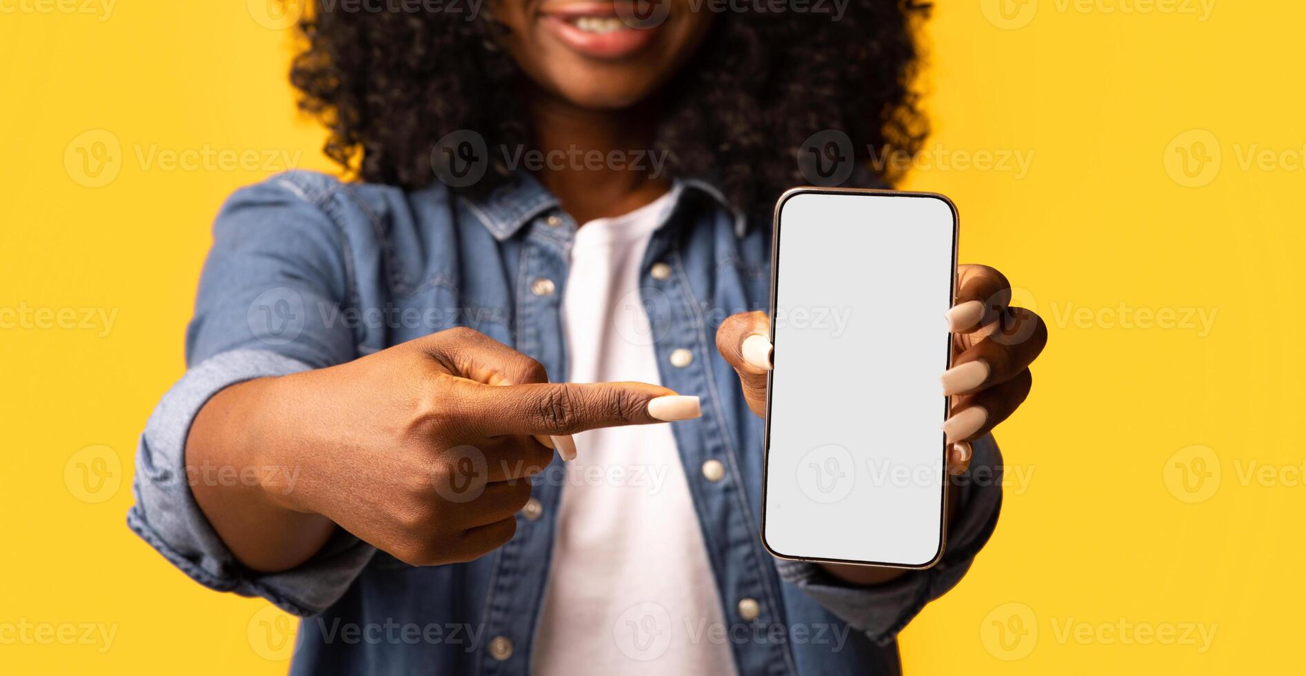 Black woman pointing at smartphone with empty screen in her hand, cropped. Unrecognizable african american lady showing modern mobile phone with blank display, closeup, mockup photo
