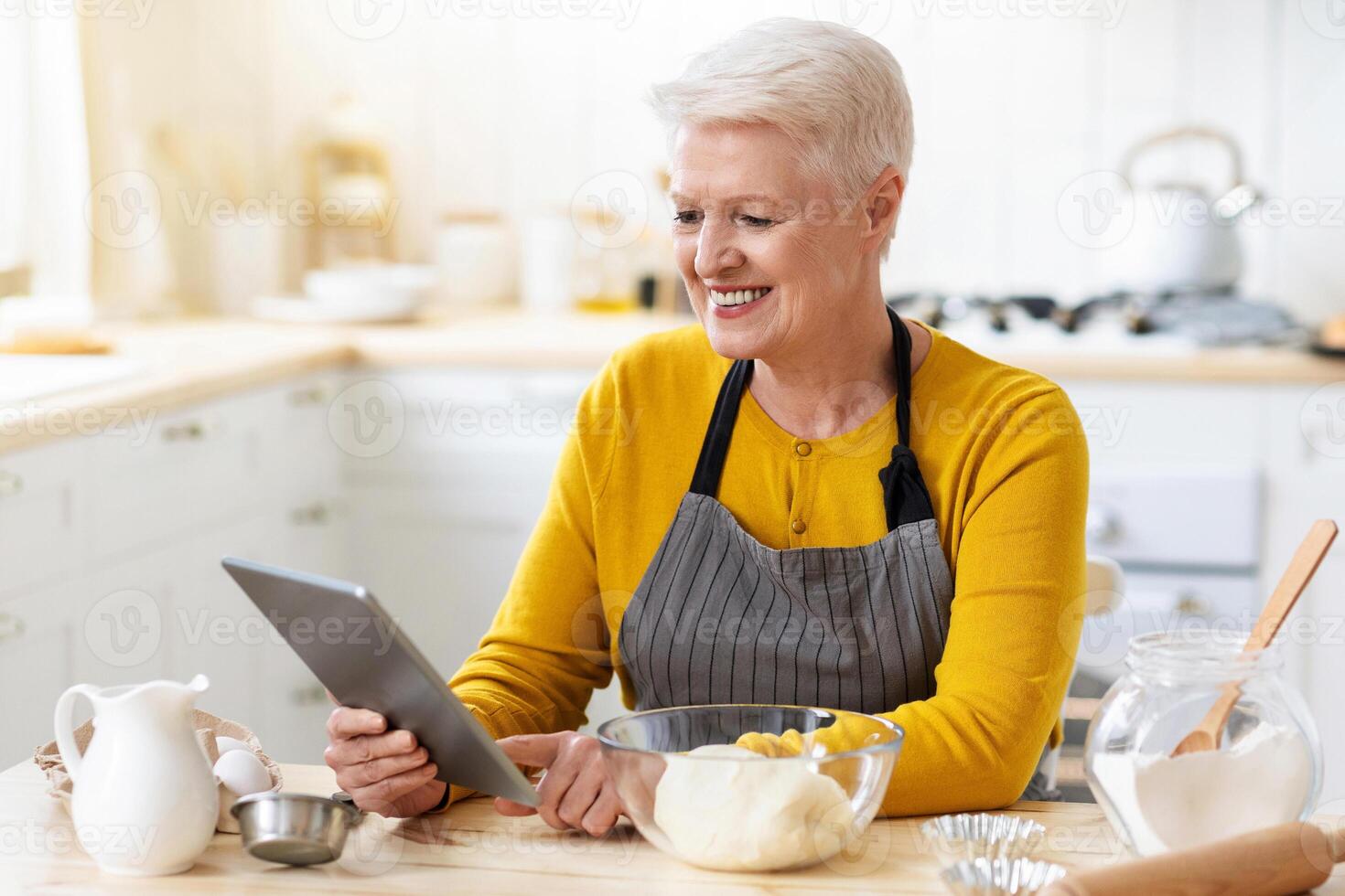 Cheerful aged lady cooking in kitchen, holding digital tablet, watching online culinary class while kneading dough, copy space. Happy old woman attending cooking webinar from home, using pad photo