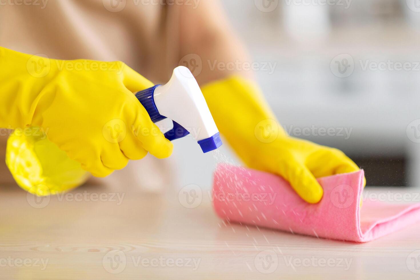 Unrecognizable maid in rubber gloves cleaning countertop at kitchen, closeup of hands with detergent and rag. Professional sanitary service worker doing house cleanup, wiping table photo