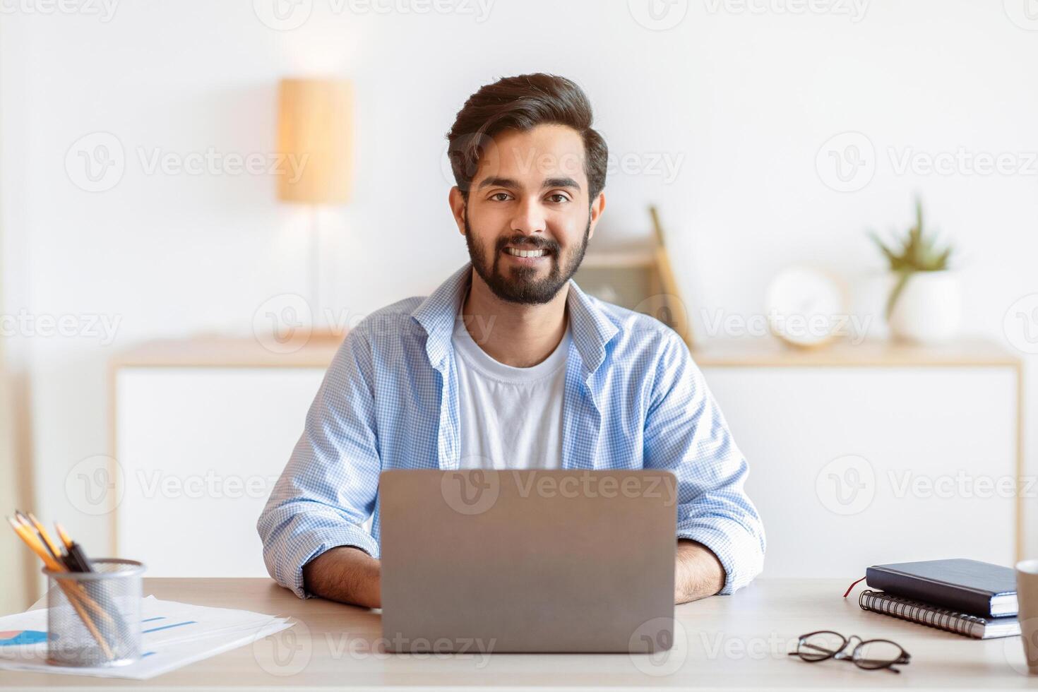Freelance Lifestyle. Portrait Of Smiling Eastern Guy Sitting At Desk With Laptop At Home Office, Handsome Arab Freelancer Man Working Remotely On Computer, Enjoying Online Distant Job, Copy Space photo
