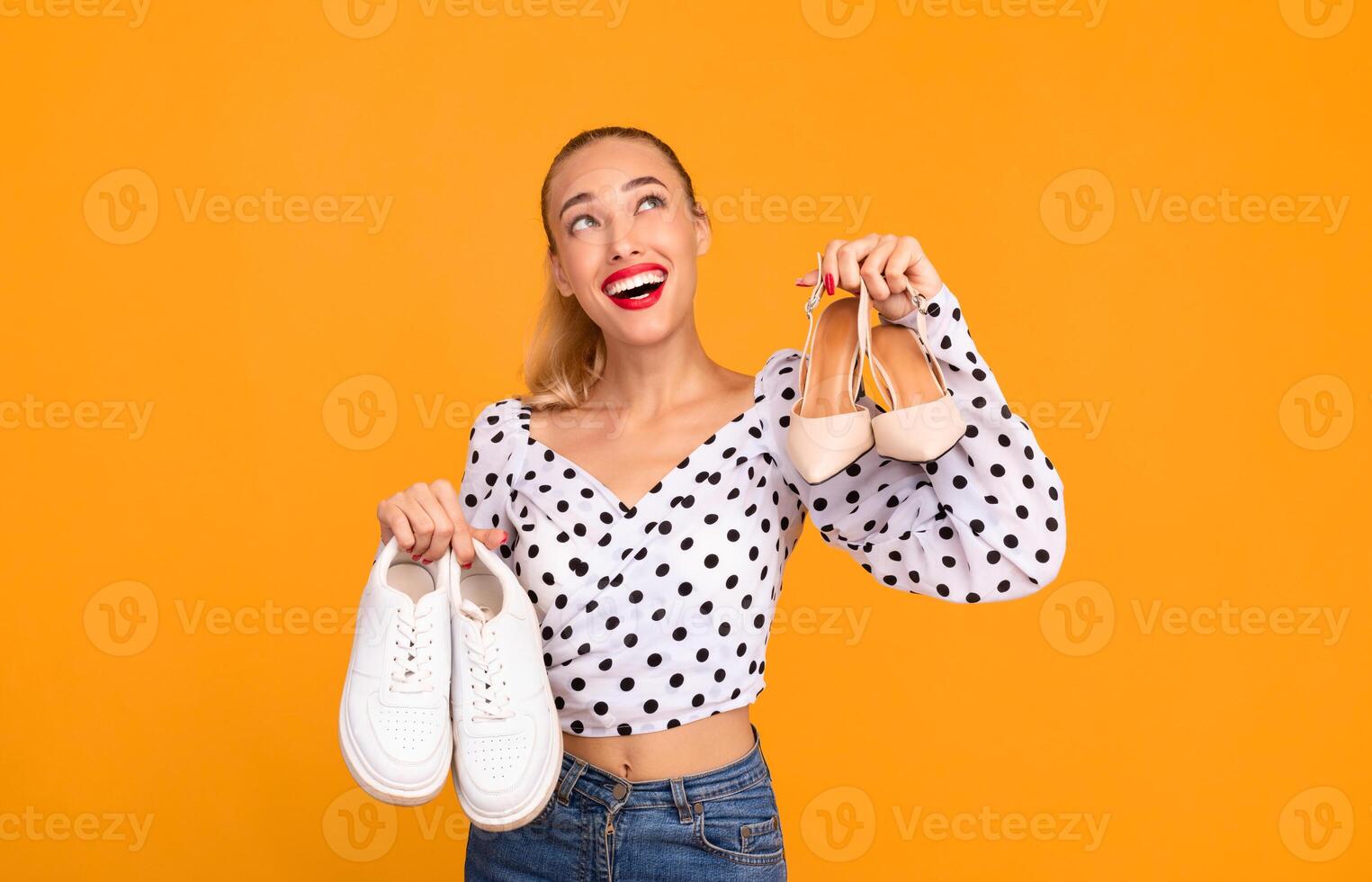 Studio portrait of a smiling stylish woman holding pair of high heels shoes and white sneakers in hands, choosing what to wear, looking up, isolated over pastel orange background wall, copy space photo