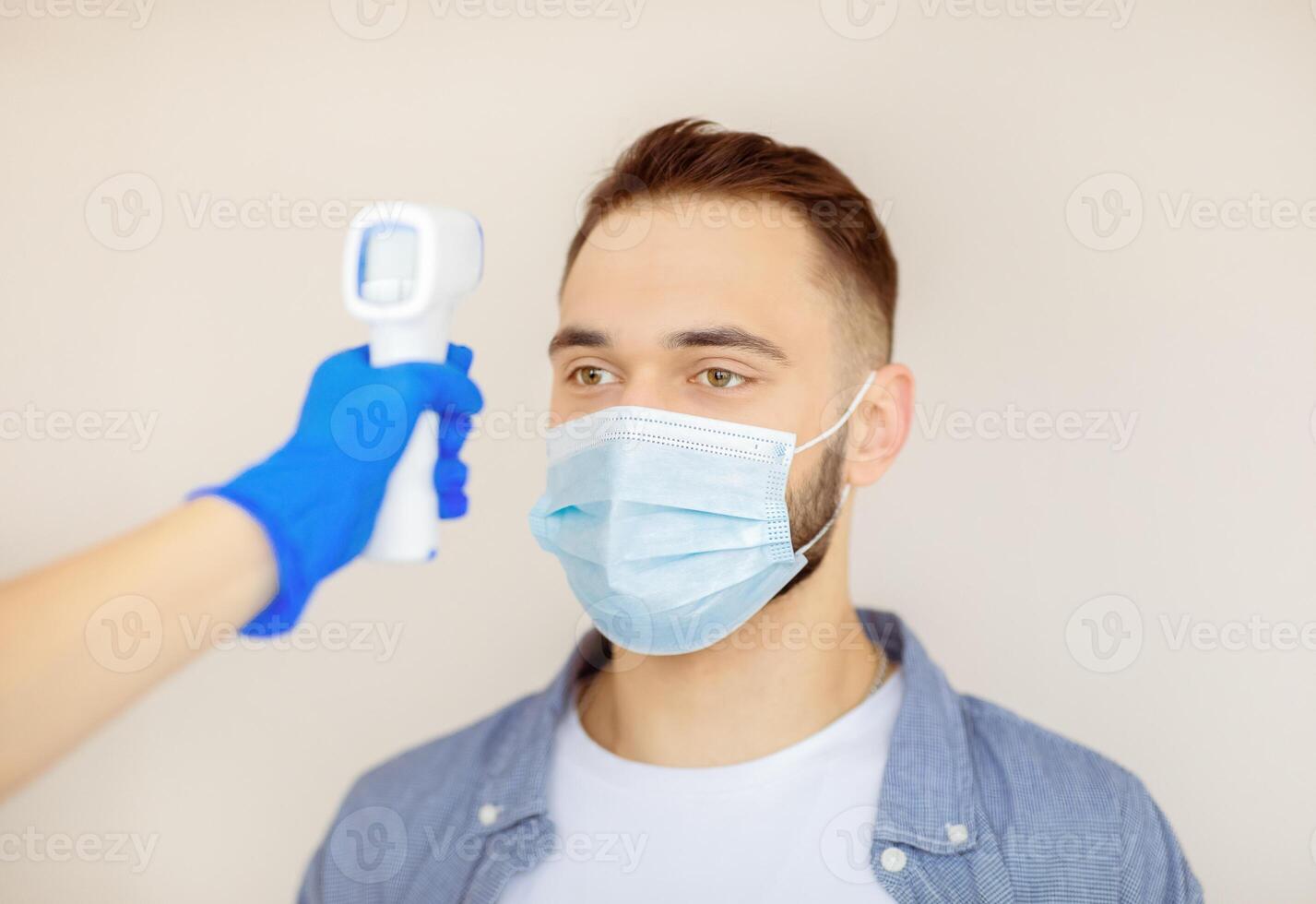 Doctor checking young man's temperature with digital thermometer before coronavirus immunization at clinic. Millennial guy in face mask visiting hospital for covid-19 vaccination photo