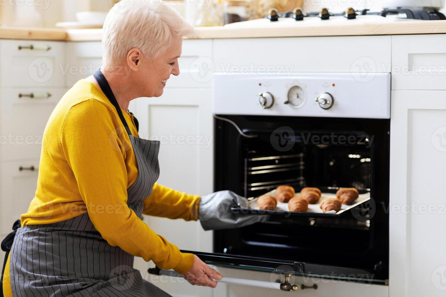 Joyful grandmother baking pastry at home, taking delicious homemade croissants out of oven, copy space. Joyful senior lady in apron opening oven, checking her pastry, cozy kitchen interior, side view photo