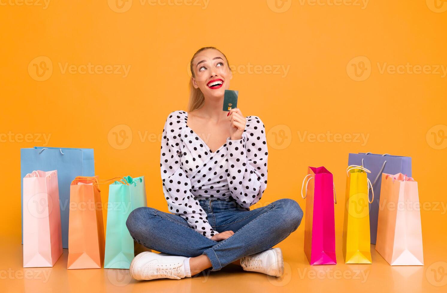 What To Buy. Portrait of trendy woman holding credit card and looking up at copy space, thinking about purchase, dreaming about new clothes, sitting on the floor at yellow studio background photo