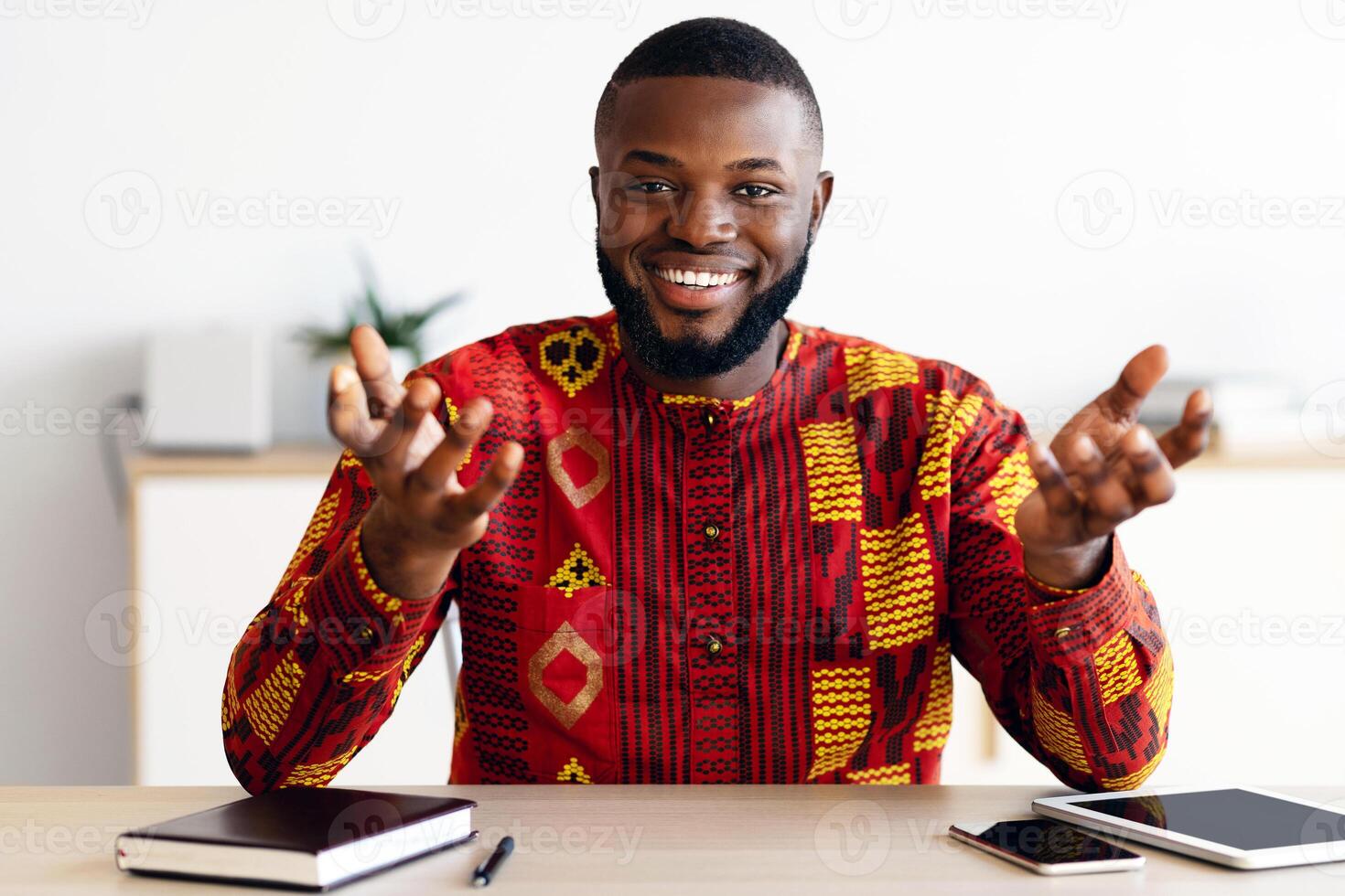 Portrait of smiling african man sitting at desk in home office, talking at camera, cheerful black advisor man in traditional clothes sharing his work experience and business ideas, free space photo