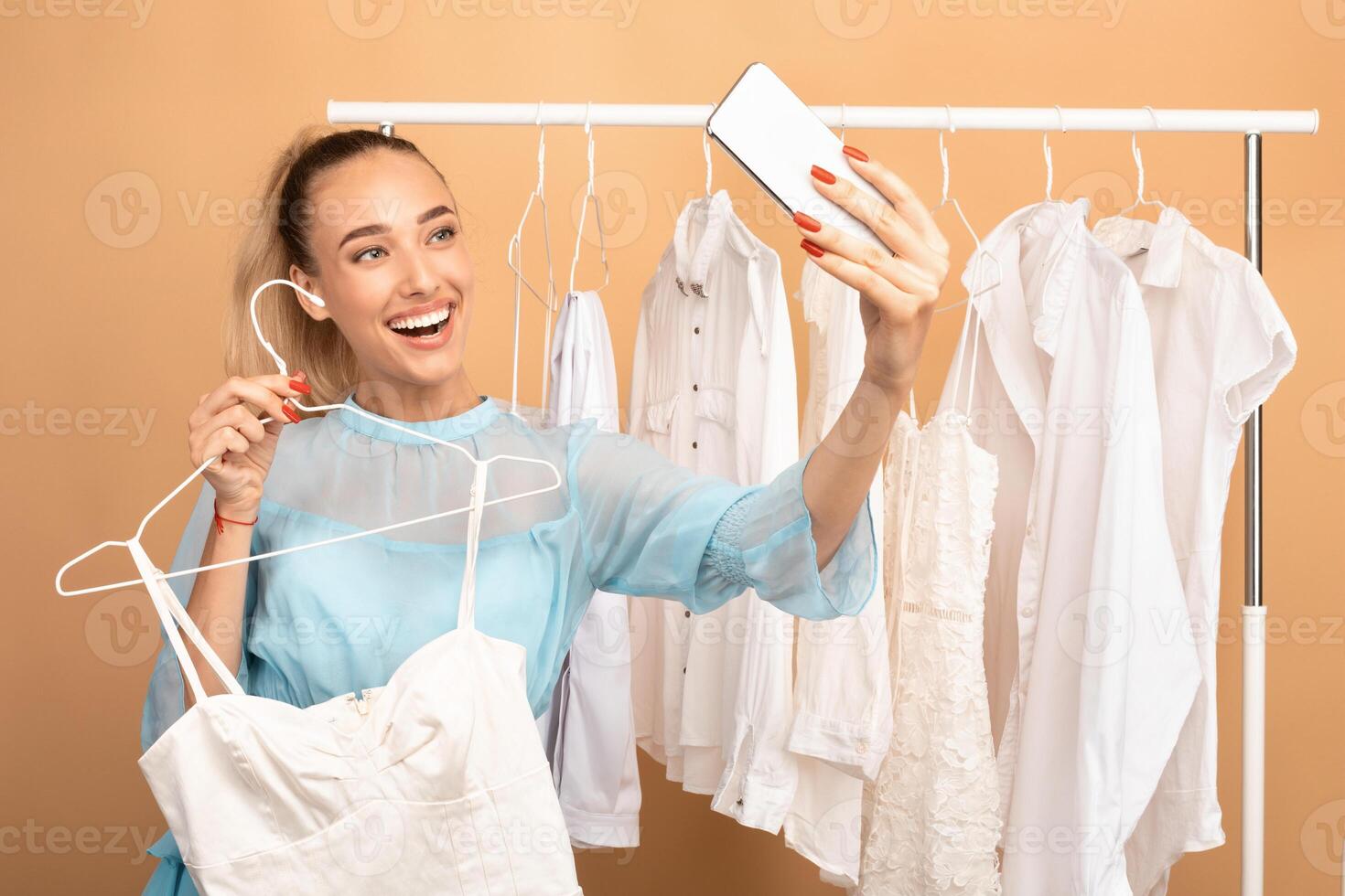 Shopping, Fashion, Style, Technology Concept. Portrait of happy laughing excited model woman taking selfie at showroom, holding clothes on hanger, smiling at gadget camera, rack in the background photo