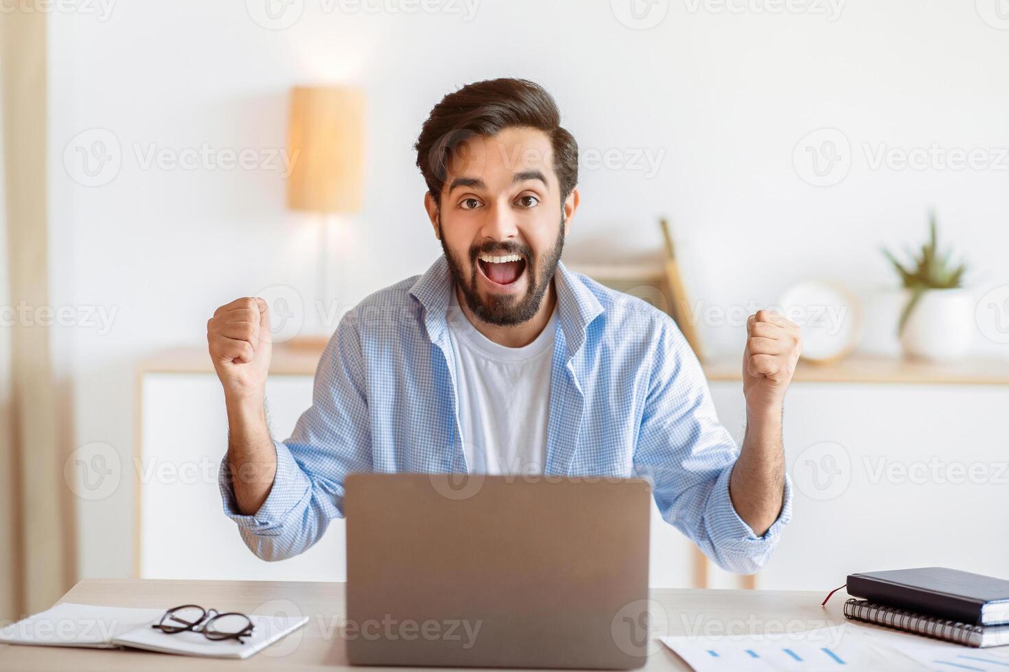 Portrait Of Overjoyed Eastern Man Celebrating Success With Laptop In Office, Excited Arab Guy Emotionally Reacting To Good News, Raising Fists And Exclaiming With Joy, Got Promotion, Copy Space photo