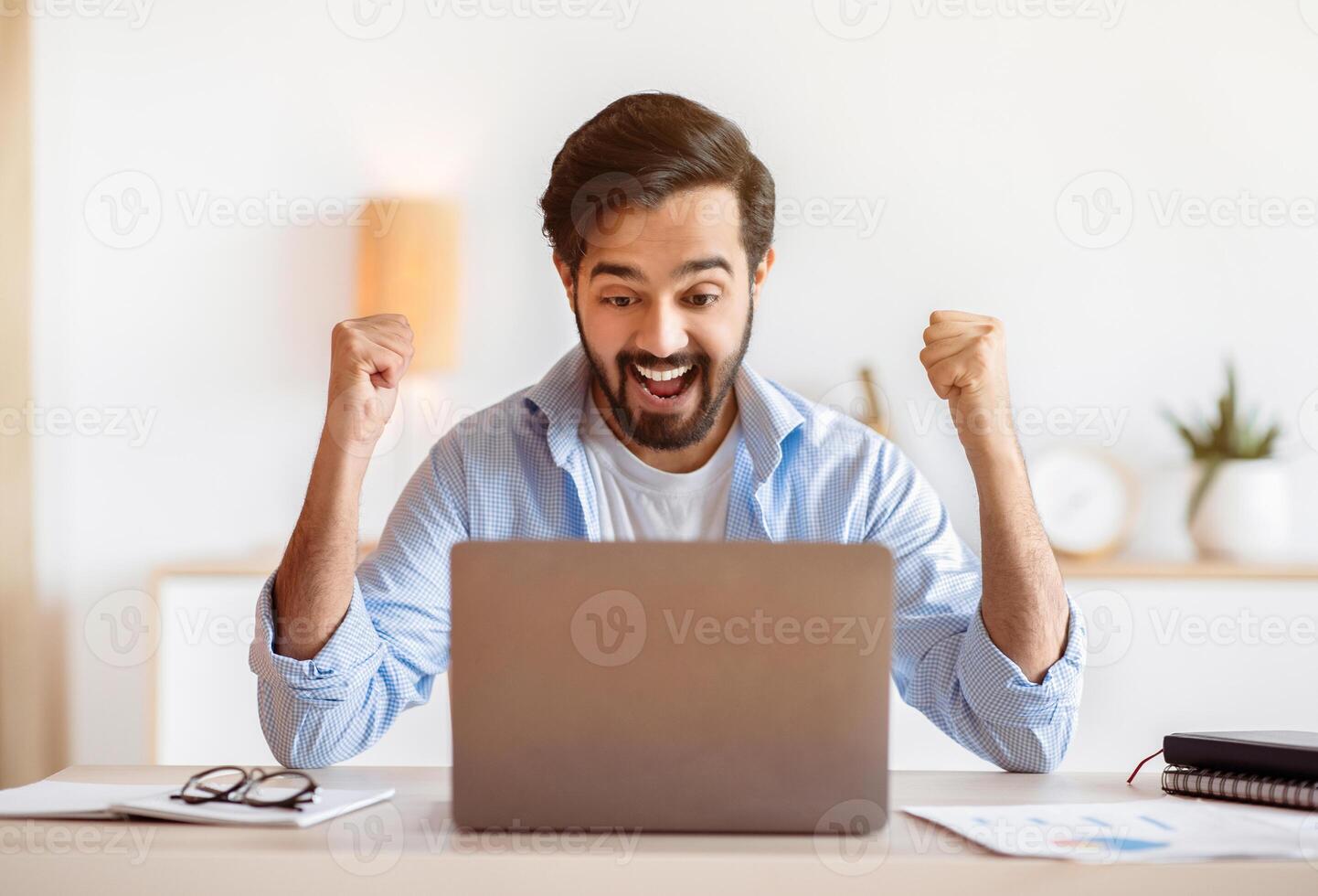 Excited arab guy celebrating success with laptop computer at home office, emotionally reacting to good news, sitting at desk, looking at computer screen, raising fists and exclaiming with excitement photo