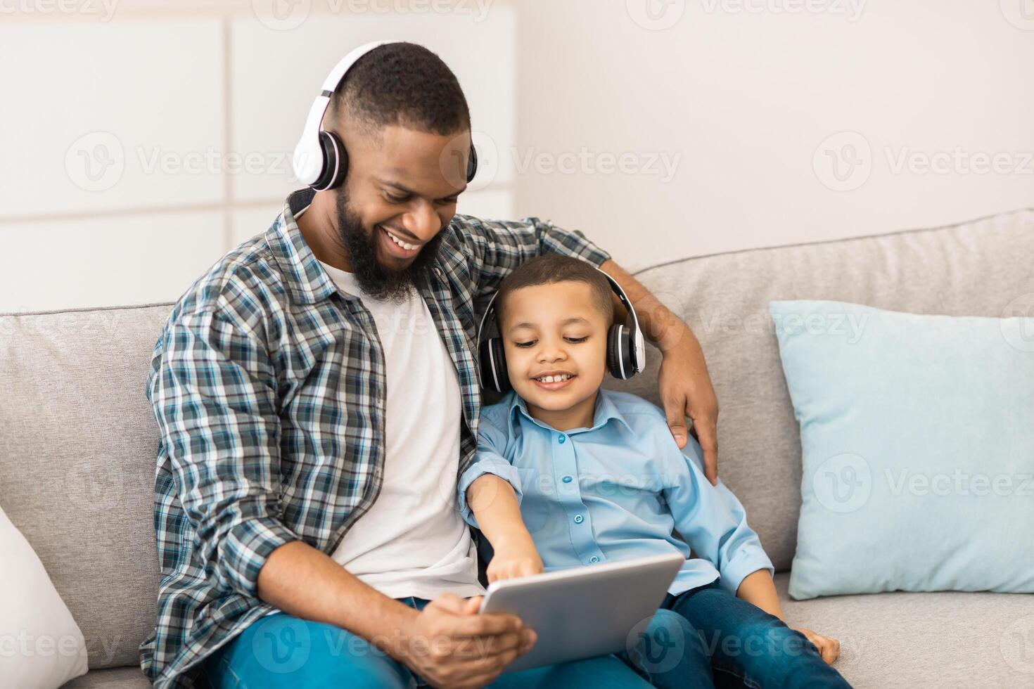 Black Father And Son Using Digital Tablet Computer Wearing Wireless Headphones Watching Movie Together Sitting On Sofa At Home. Weekend Leisure. Family, Gadgets And Modern Lifestyle Concept photo