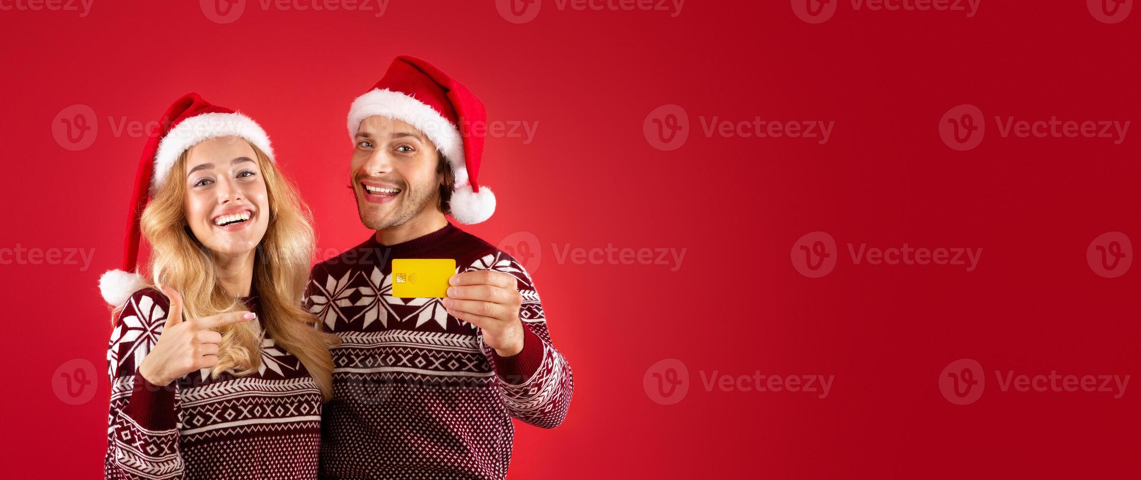 Xmas shopping concept. Affectionate young couple in Santa hats pointing at credit card on red background photo