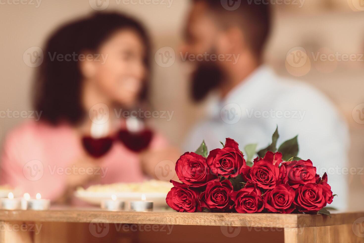 Bonding Concept. Bouquet of flowers lying on table, selective focus on red roses. Happy black man and woman spending time together at home or cafe, having dinner, drinking wine in blurred background photo