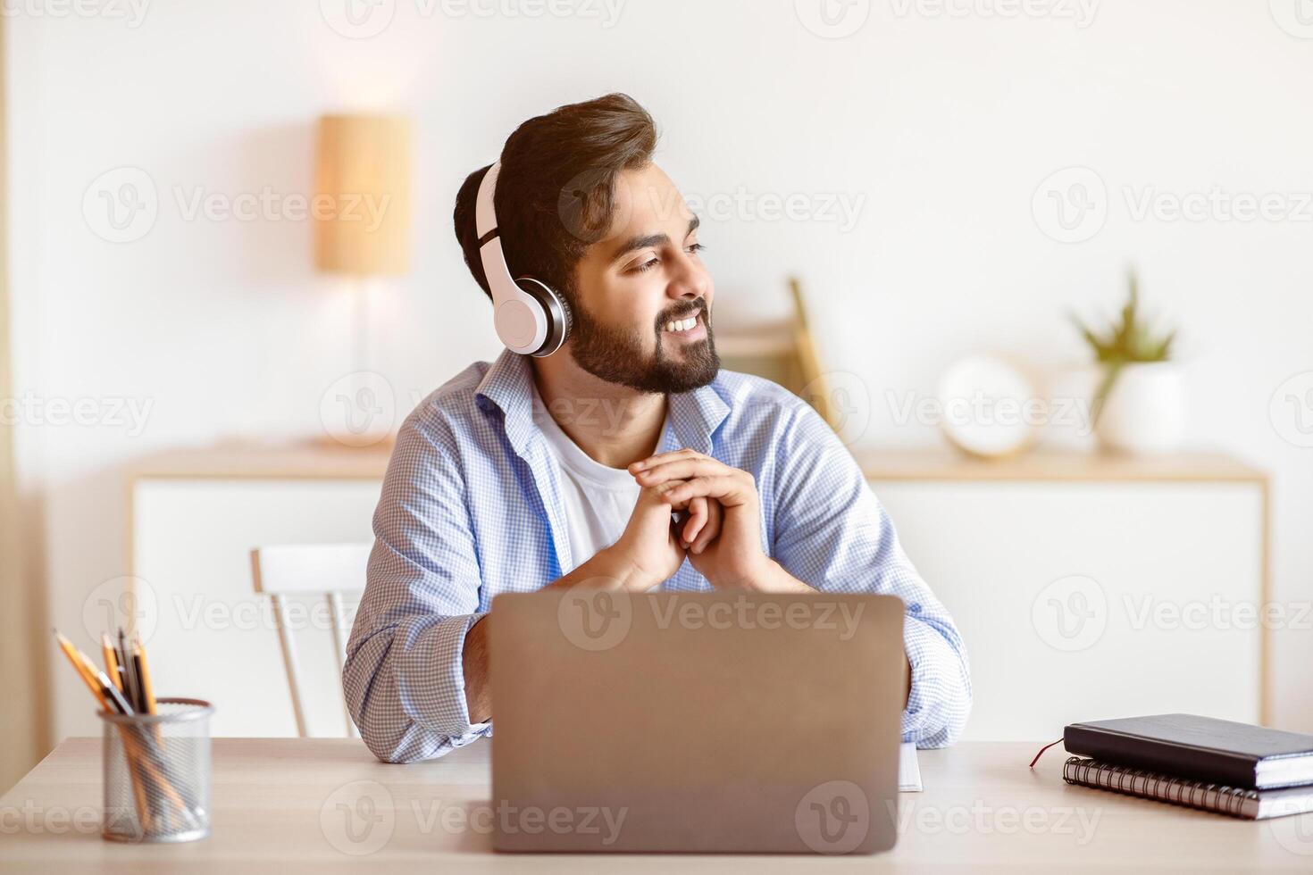 Break In Work. Smiling arab man listening music in headphones while working on laptop at home office, relaxed eastern guy sitting at desk with computer and looking aside, thinking about something photo