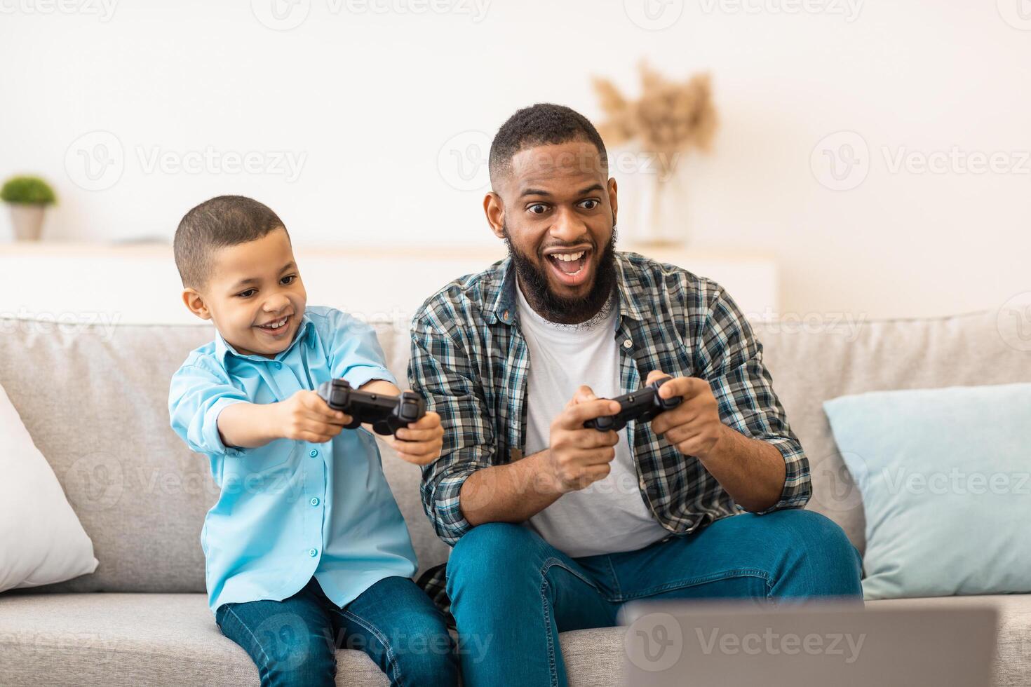Excited African Daddy And His Son Boy Playing Game Bonding And Having Fun Together Sitting On Sofa In Living Room At Home. Family Leisure And Entertainment. Weekend With Father photo