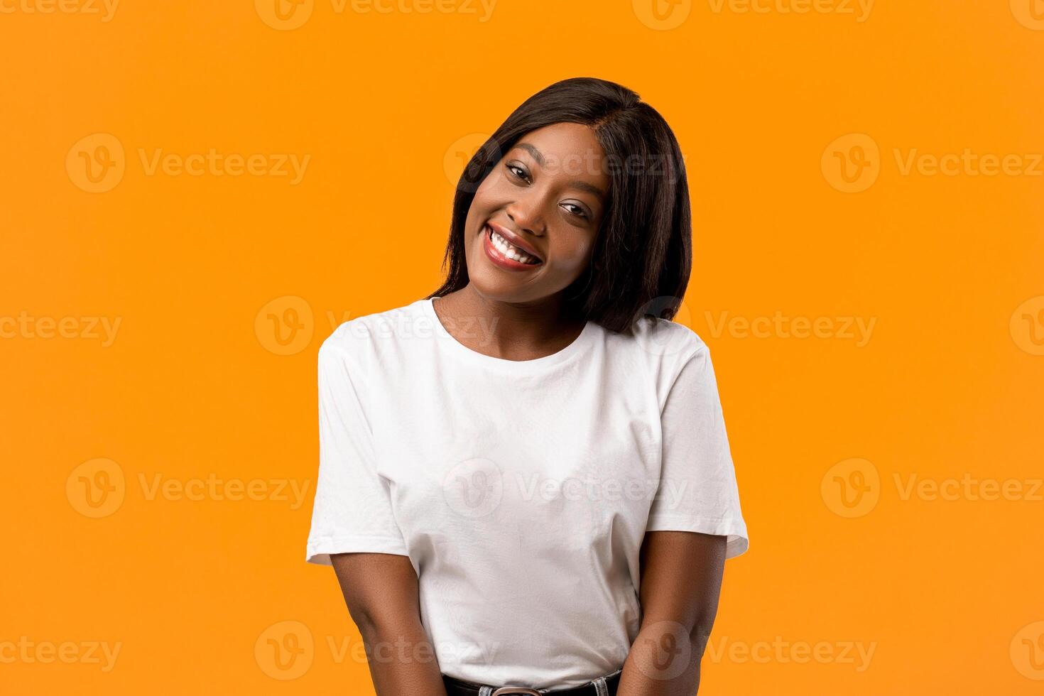Cute cheerful black young lady in white t-shirt posing on orange studio background, copy space. Beautiful african american woman with shining fair hair smiling at camera, flirting photo
