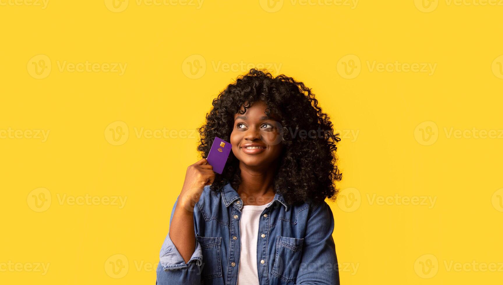 Cheerful african american young woman with curly hair holding credit card and looking at copy space over yellow studio background, panorama. Banking, loan, unlimited shopping concept photo