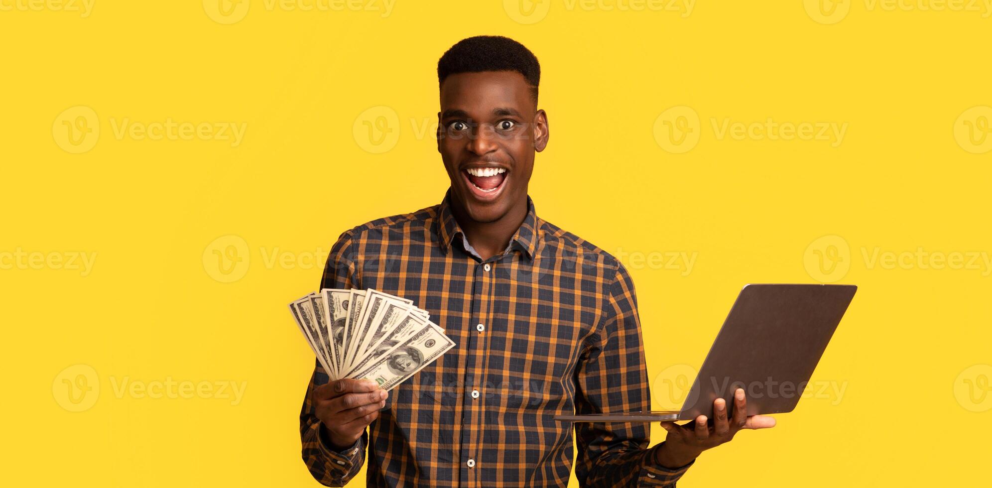Online Lottery. Excited black guy with dollar cash and laptop celebrating success, overjoyed african american man holding money and computer, standing isolated over yellow background, free space photo