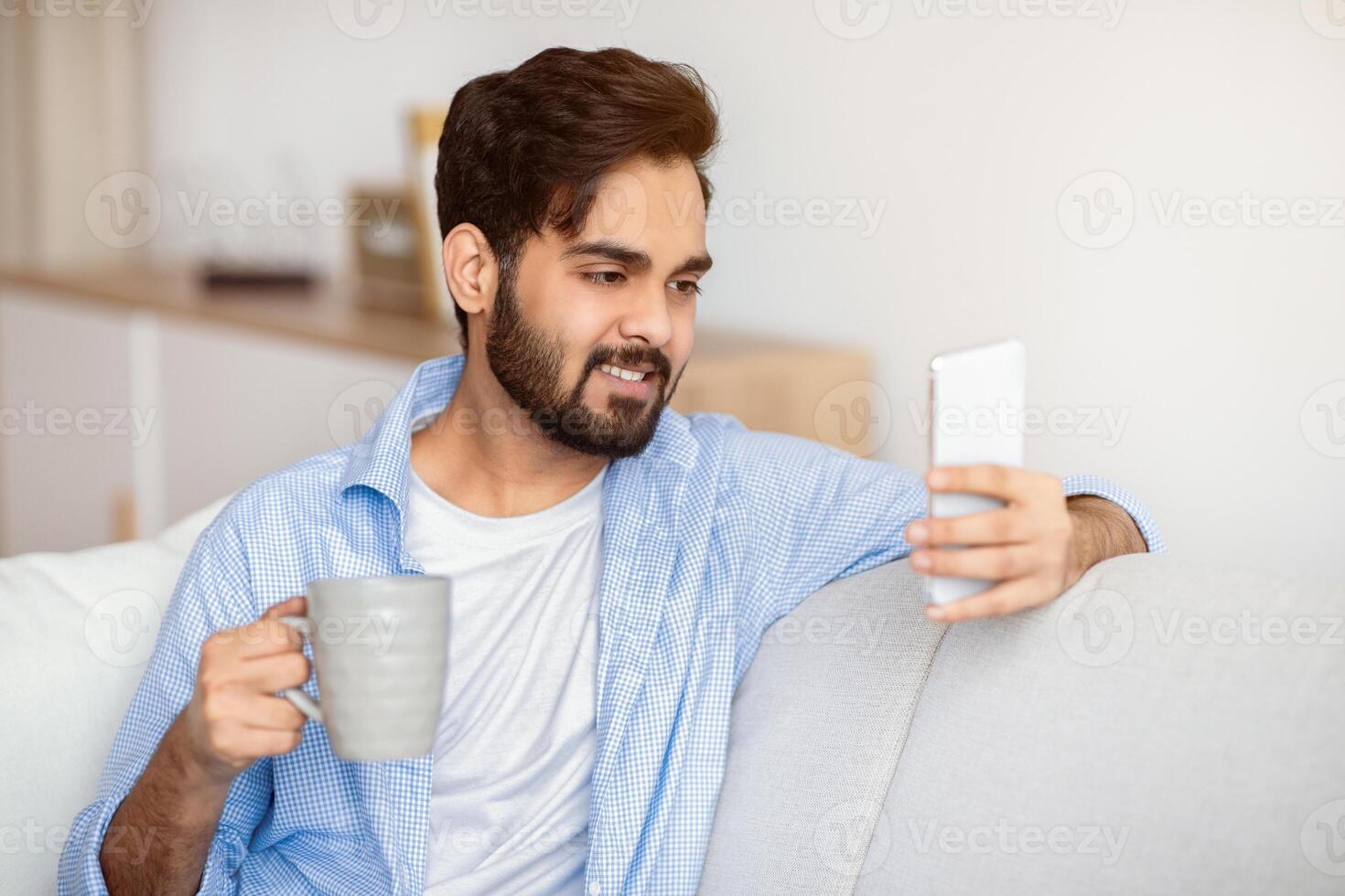 Domestic Leisure. Young Eastern Man Using Smartphone And Drinking Coffee At Home, Watching Online Videos On Cellphone And Enjoying Hot Drink While Relaxing In Living Room, Closeup Shot photo