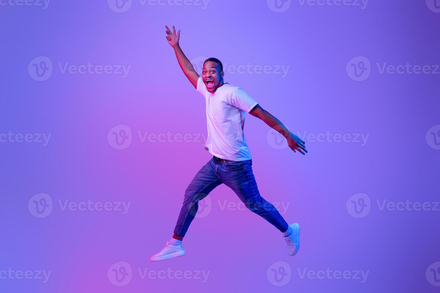 Excited Funny Black Guy Jumping Up In Neon Light Over Purple Background, Full-Length Portrait In Motion Of Euphoric African American Man Jump In The Air With Open Mouth And Raised Hands, Copy Space photo