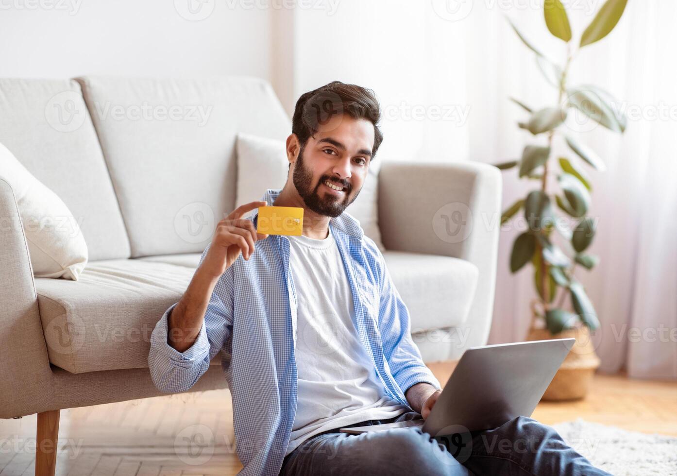 Smiling Arab Man Using Laptop Computer At Home And Demonstrating Credit Card At Camera, Millennial Eastern Guy Enjoying Online Shopping Or Easy Payments While Resting On Floor In Living Room photo