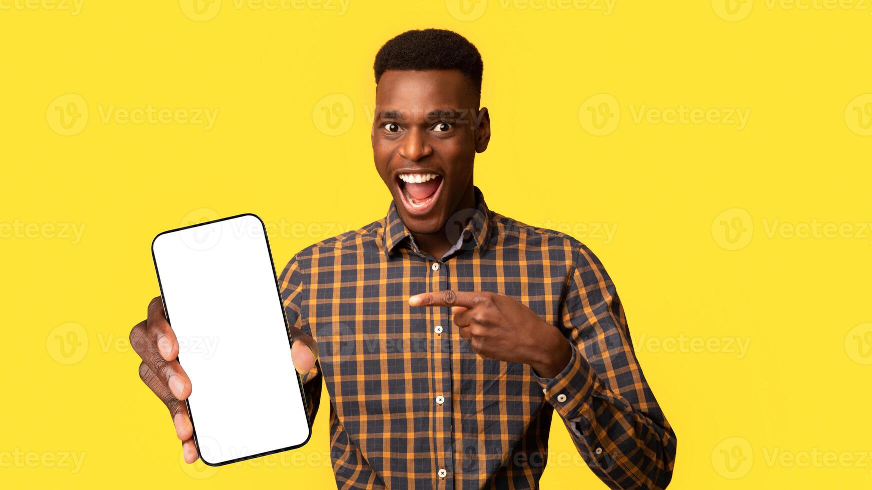 Mobile Offer. Excited Black Guy Pointing At Smartphone In His Hands, Emotionally Reacting To New App, Overjoyed Millennial African American Man Standing Isolated Over Yellow Background, Copy Space photo